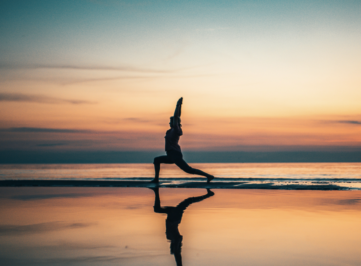 Person practicing yoga in a warrior pose on the beach at sunset with a reflection on the wet sand.