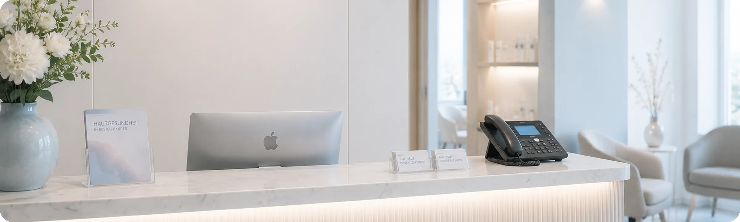 Modern reception desk with Mac computer, phone, flower vase, and brochure in a bright waiting area.