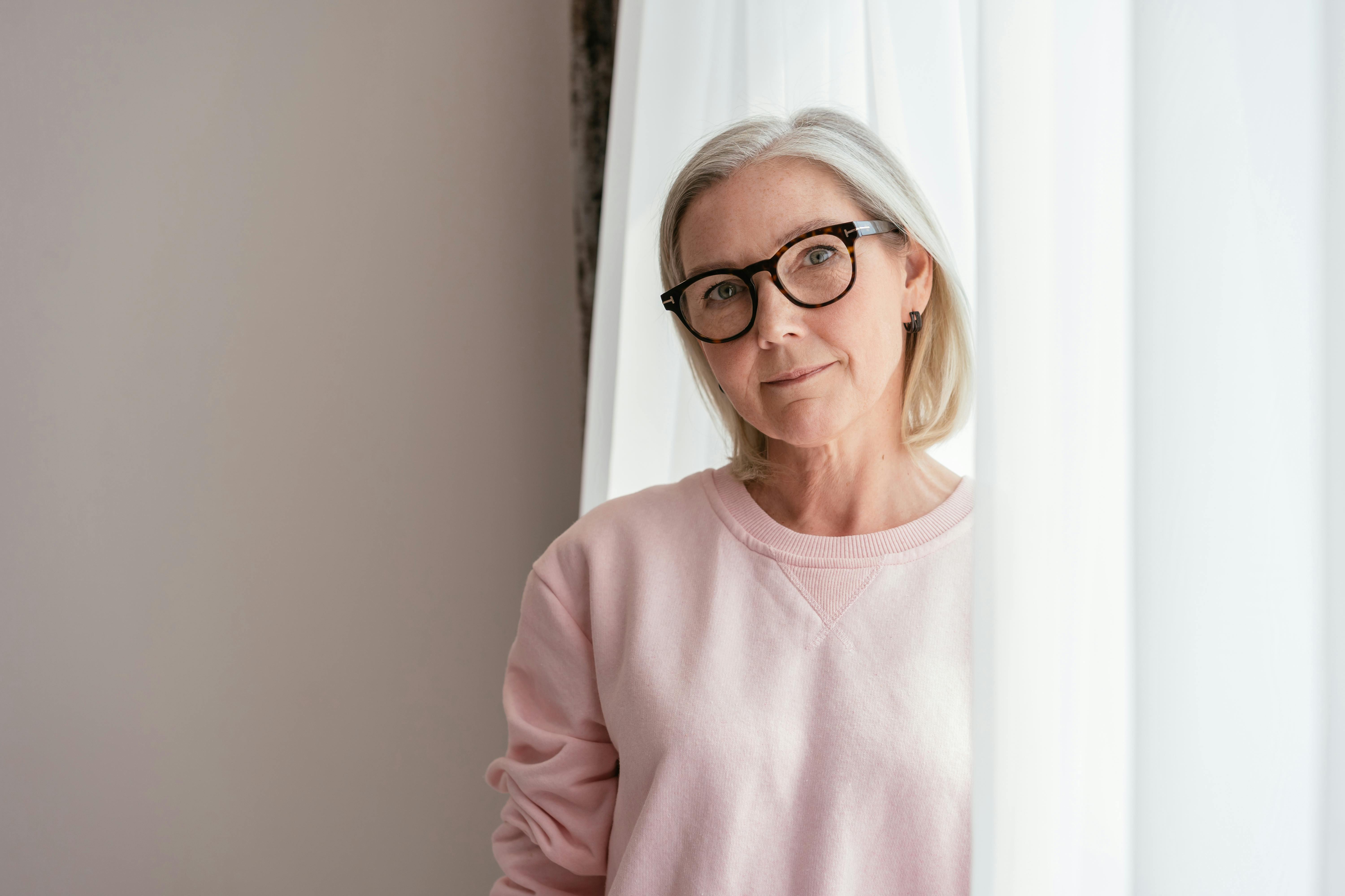 Middle-aged woman with short gray hair and glasses wearing a light pink sweater, standing by a white curtain.