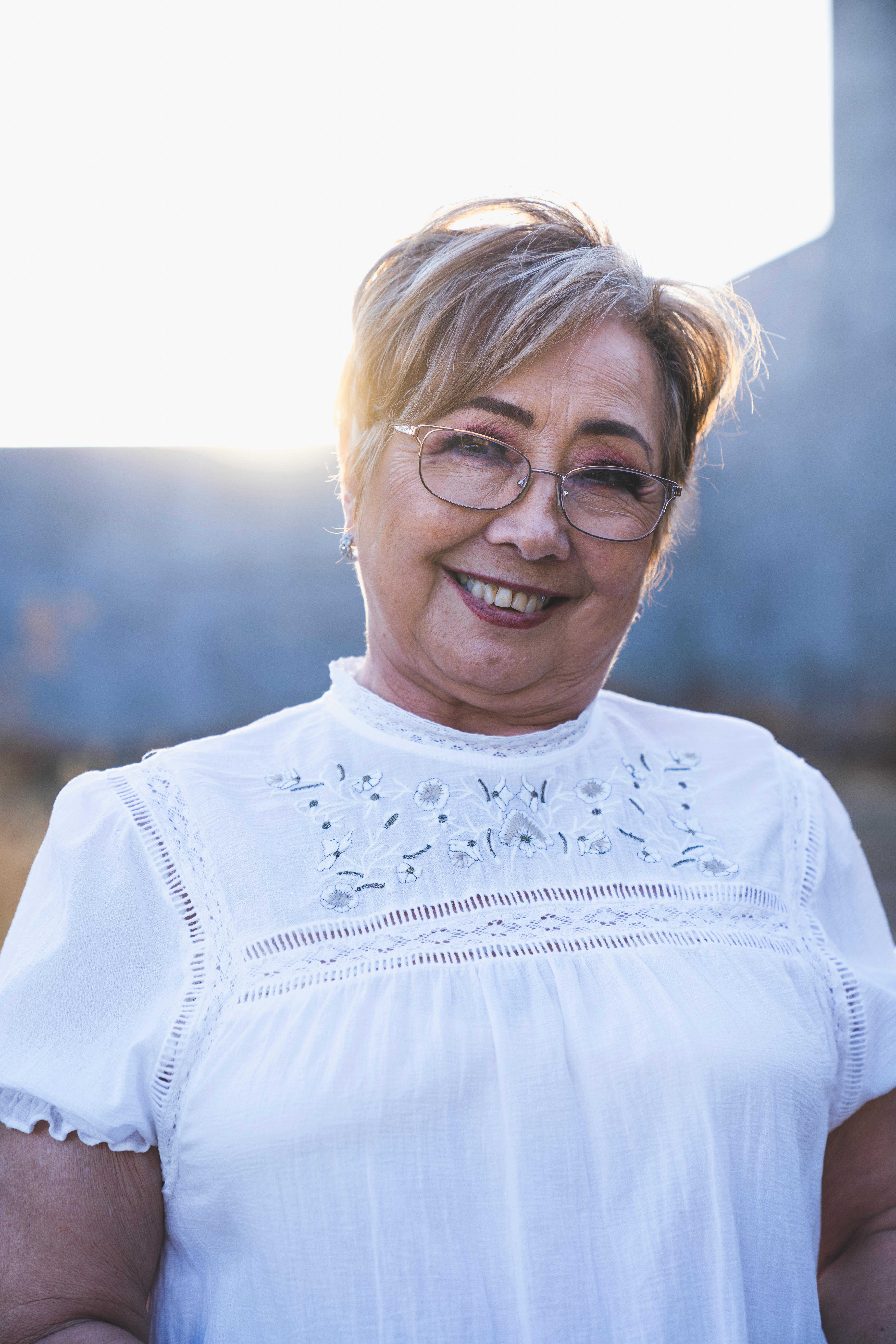 Smiling older woman with short hair and glasses wearing a white embroidered blouse outdoors with sunlit background.