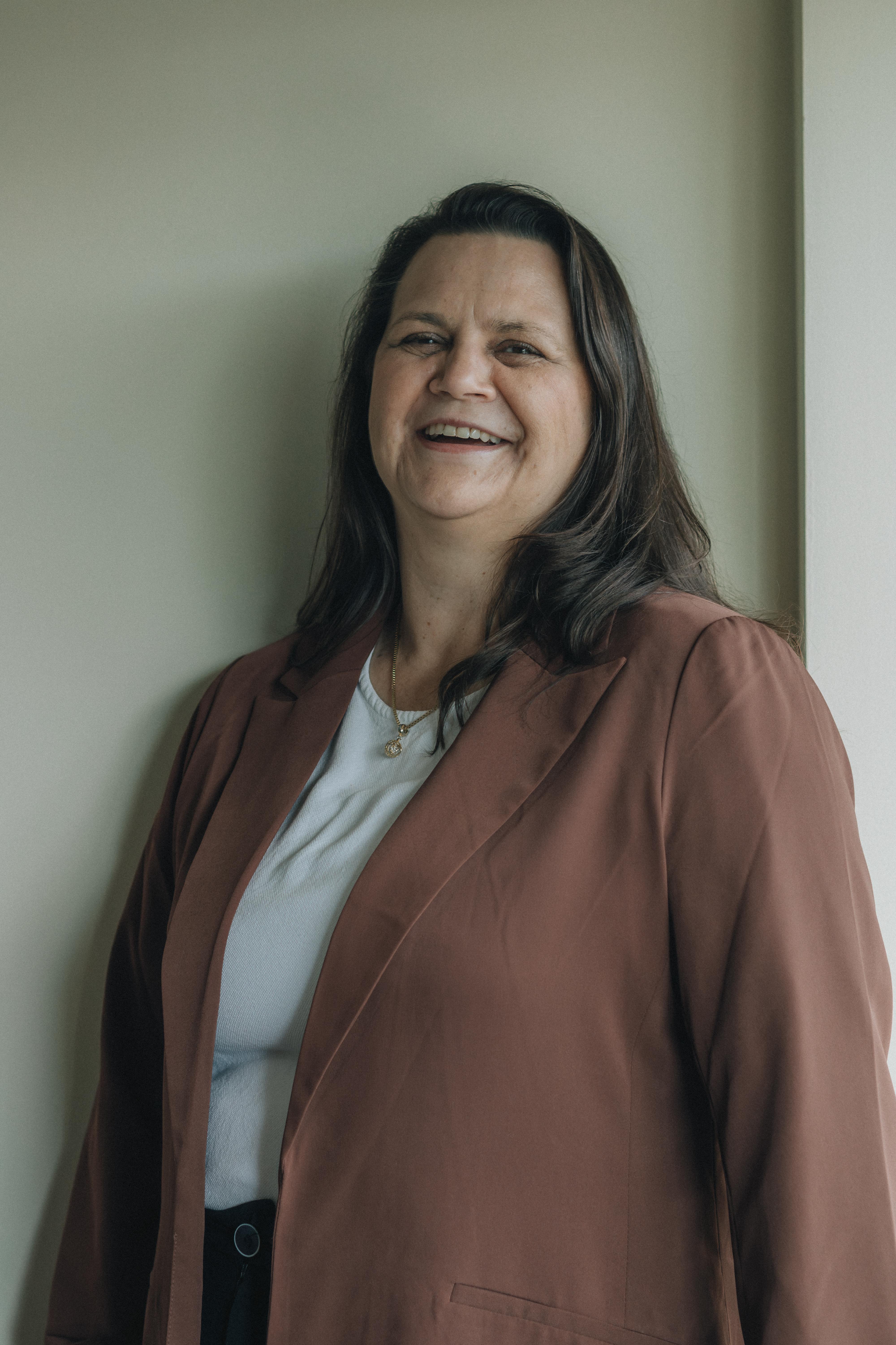 Smiling woman with long dark hair wearing a brown blazer and white top standing against a light wall.