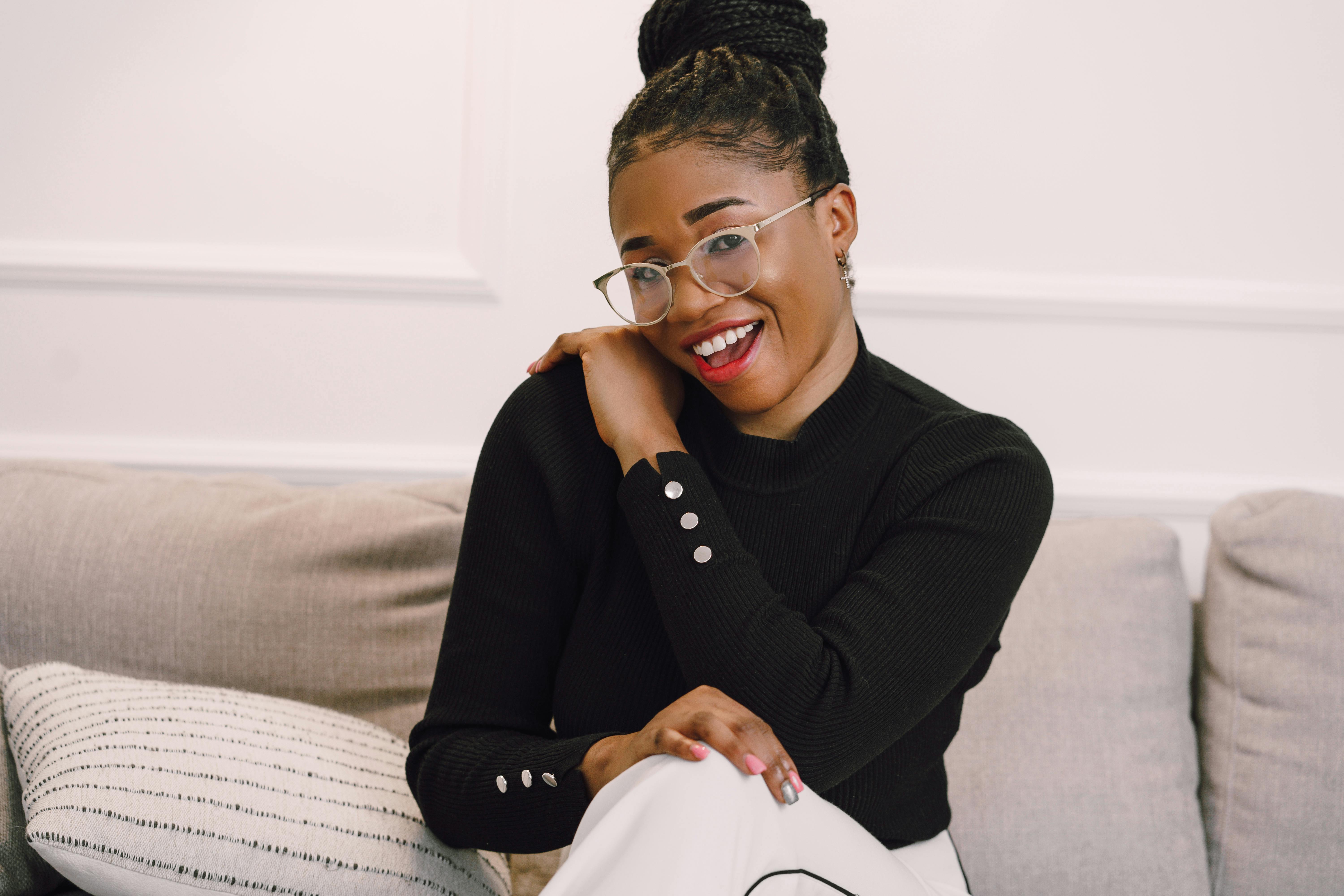 Smiling woman with braided hair in a black sweater and glasses sitting on a beige couch with a striped pillow.