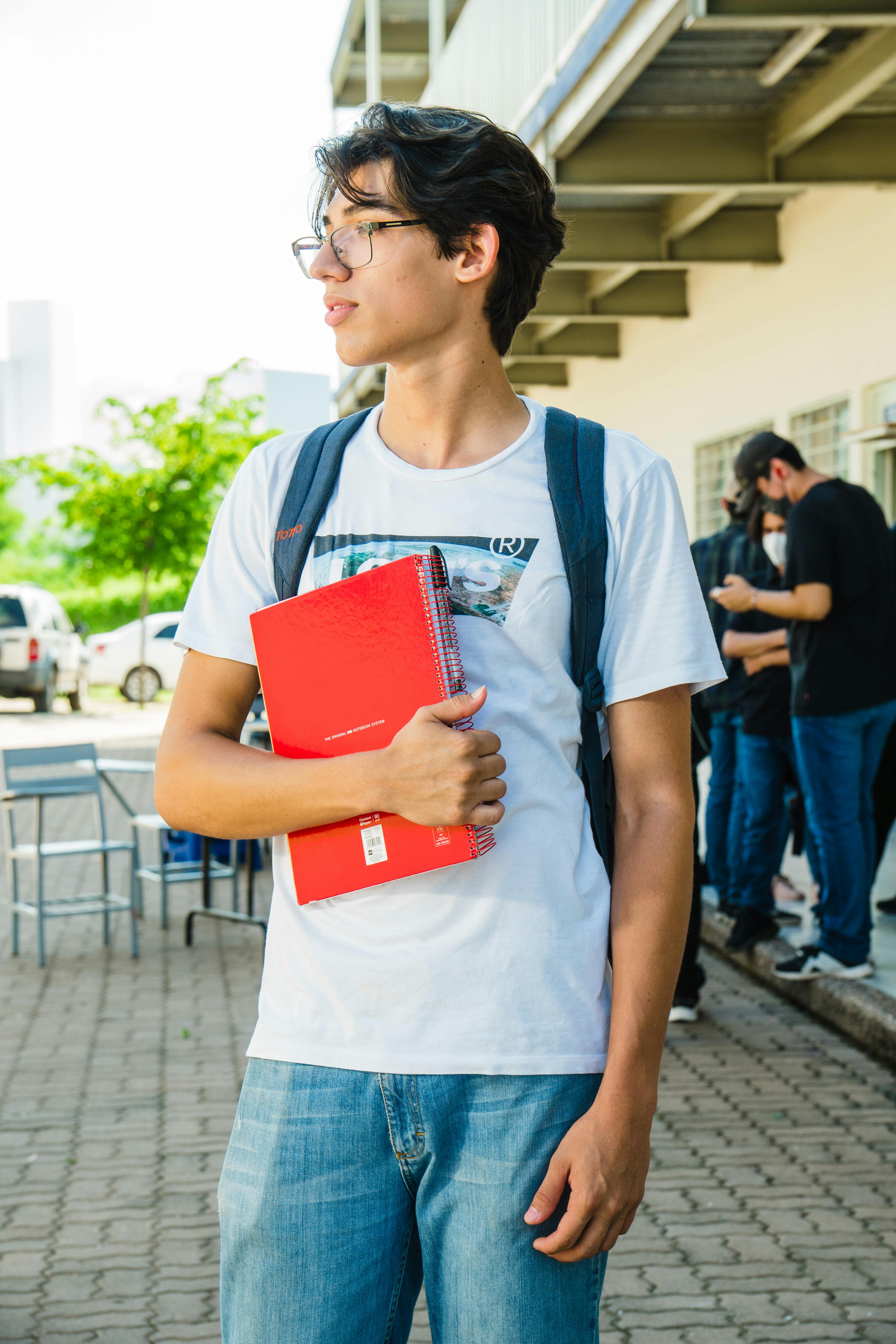 Young male student in glasses holding a red notebook and wearing a white t-shirt and backpack outdoors near a building.