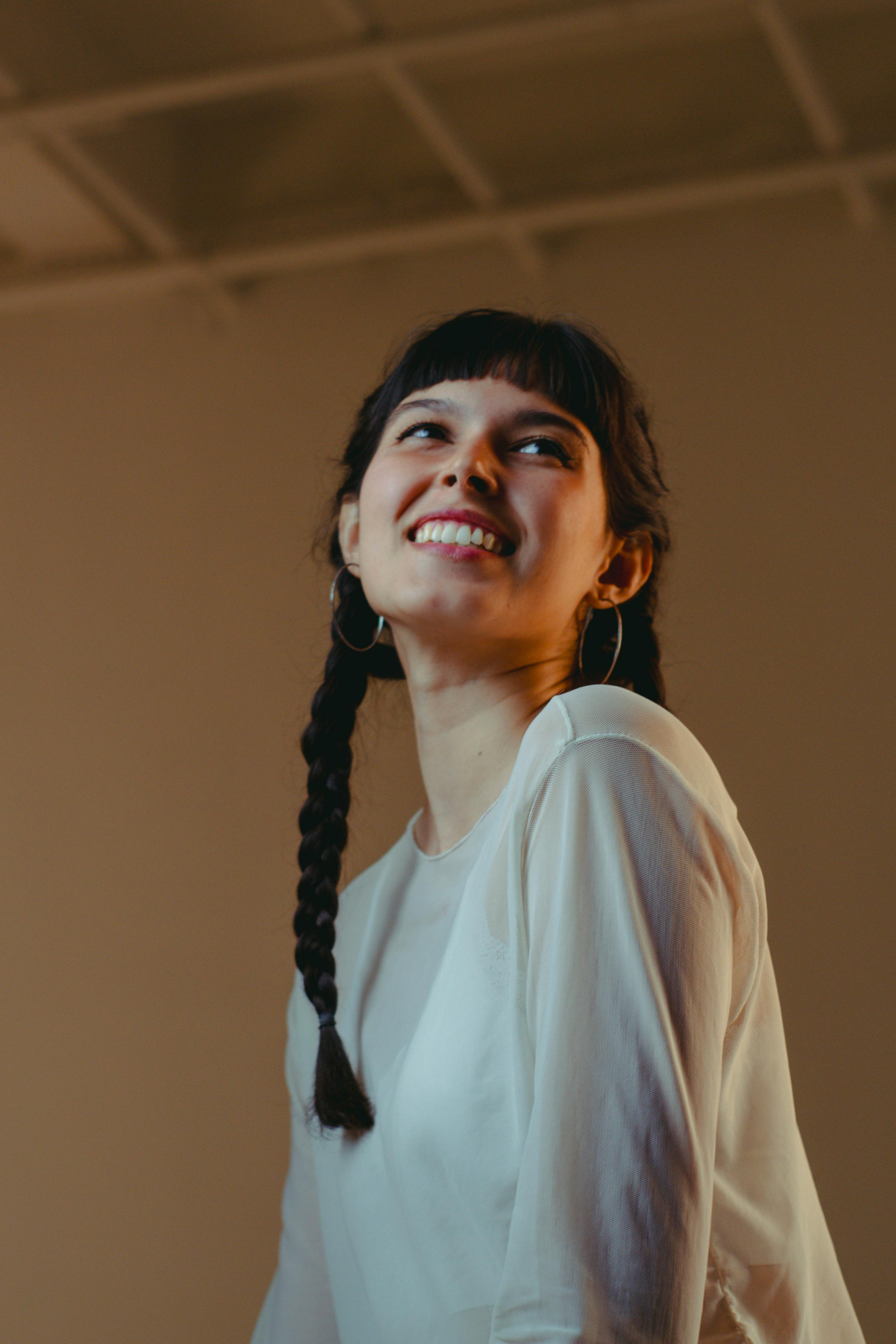 Smiling woman with braided hair and hoop earrings wearing a white sheer top looking upward.