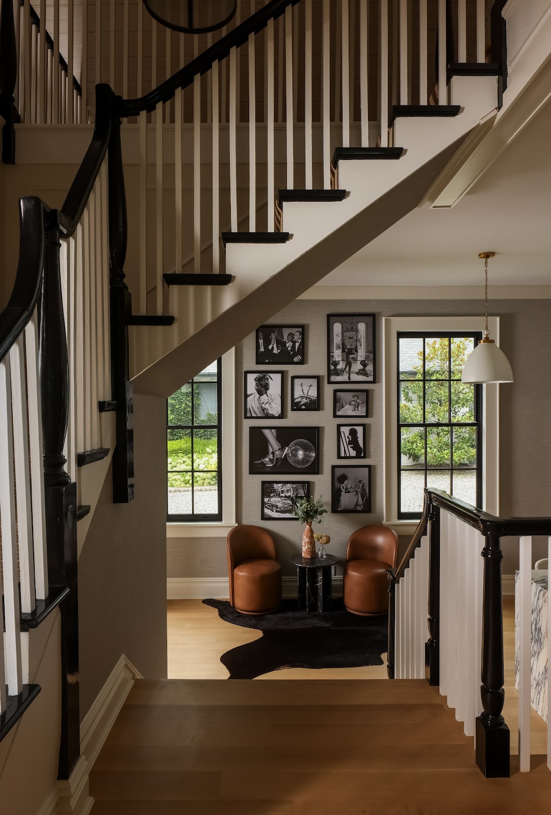 View through a dark wood staircase railing into a sitting nook with leather chairs, a black-and-white gallery wall, and original iron-grid windows in a renovated 1907 Long Island carriage house.