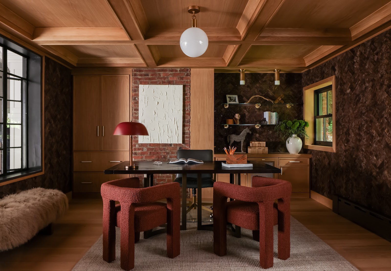 Home office with a custom white oak coffered ceiling, exposed brick, dark wallcovering, warm wood cabinetry, and two burnt orange boucle chairs around a deep brown wood desk.