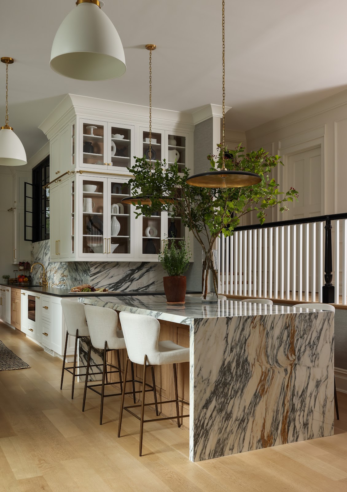 Close view of an Arabescato Antico marble kitchen island with white upholstered bar stools, glass-front cabinetry, and brass pendant lights in a historic carriage house renovation.