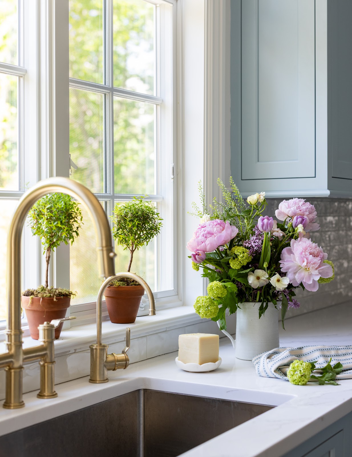 Close-up of a kitchen sink with a brass bridge faucet, potted topiaries on the windowsill, pink peonies in a white pitcher, and white marble countertops.