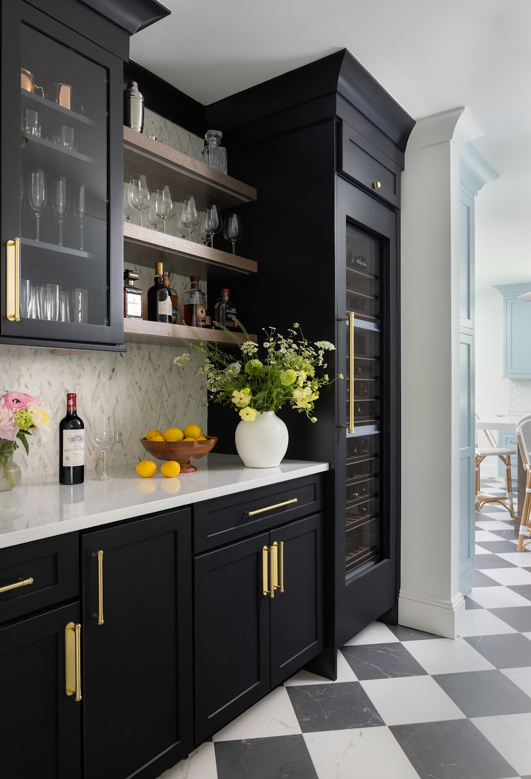 Bar area with matte black cabinetry, brass hardware, cerused oak open shelves with glassware and spirits, herringbone marble backsplash, and a wine refrigerator.