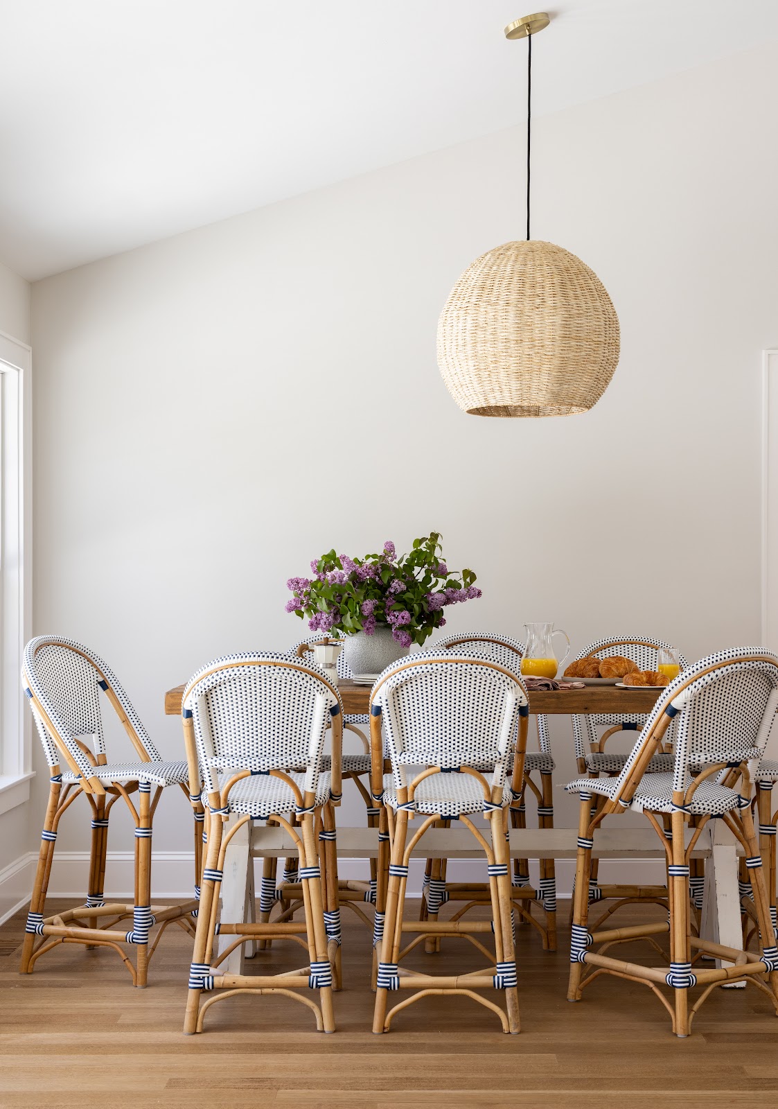A wood dining table surrounded by navy and white rattan bistro chairs, set with lilacs, orange juice, and croissants beneath an oversized woven pendant light.