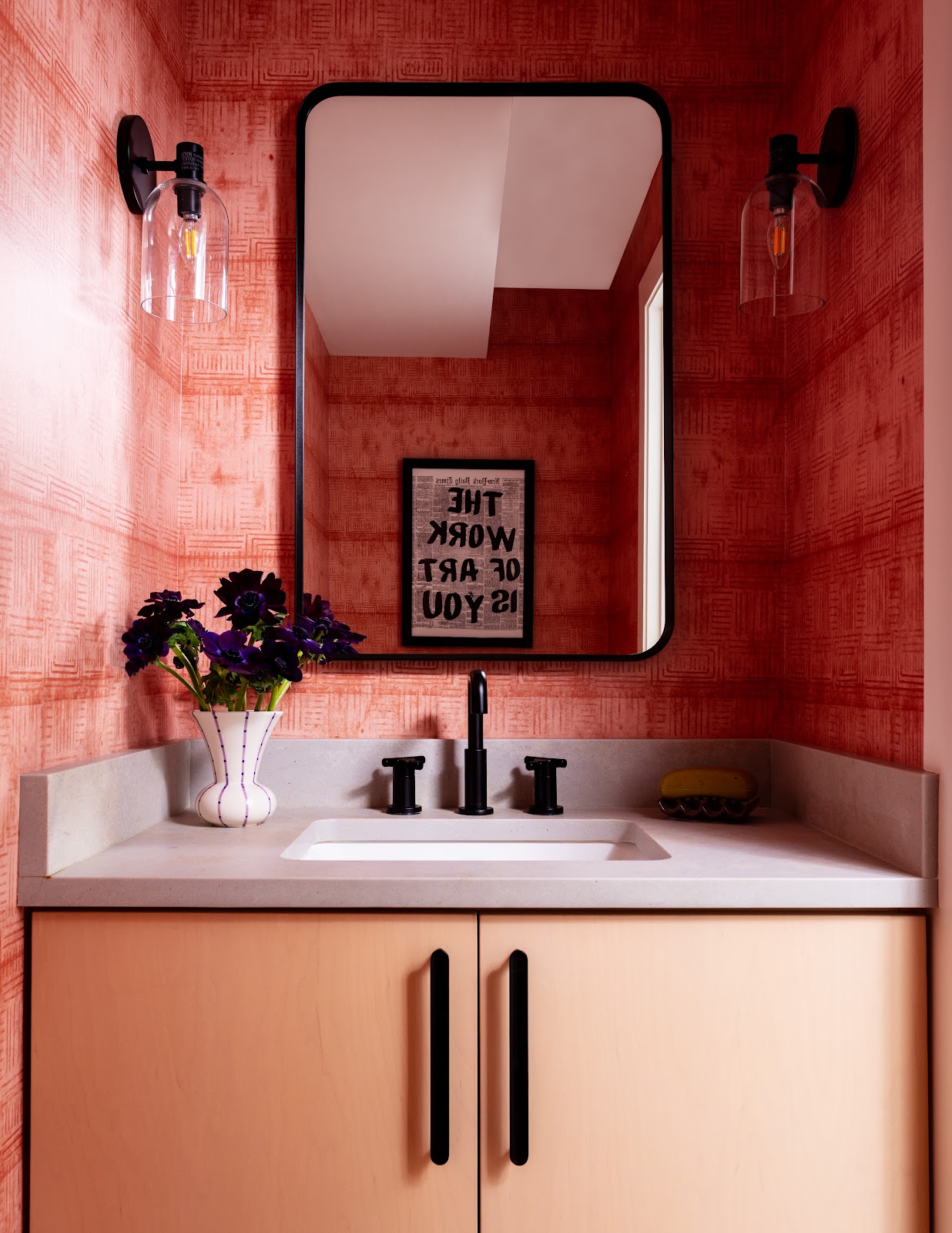 Powder room with deep coral patterned wallpaper, light wood vanity, matte black fixtures, and globe sconces in a renovated Long Island beach house.