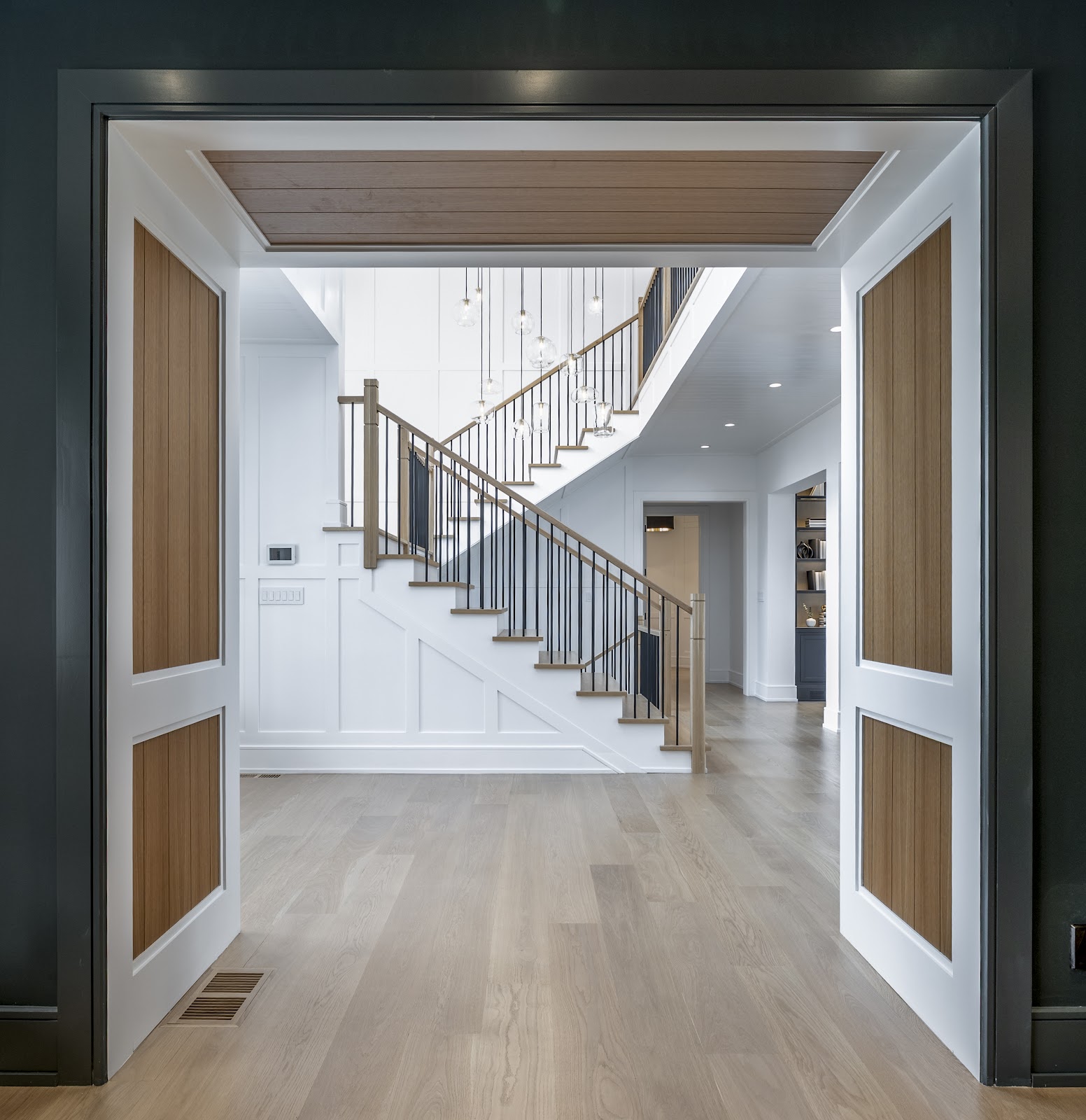 View through dark-framed double entry doors into the foyer of a contemporary Lloyd Harbor home, with white oak staircase and glass pendant lights visible.