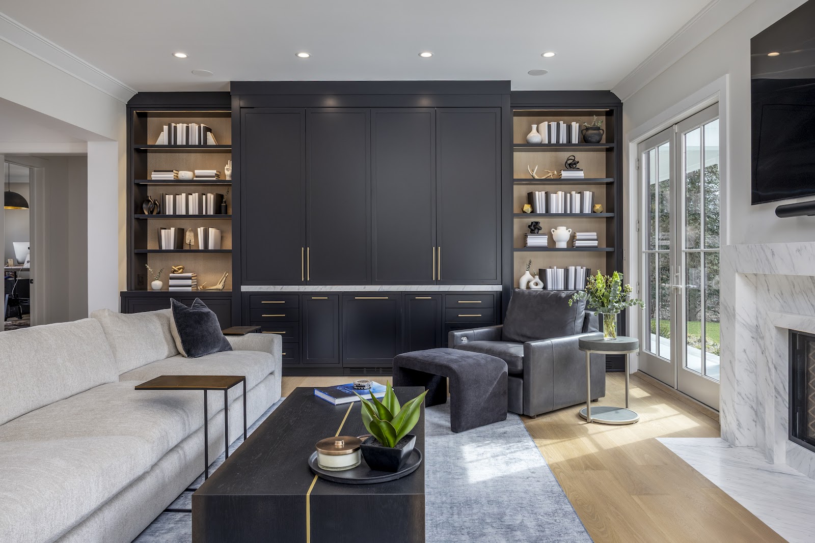 Living room with a full wall of dark charcoal built-in cabinetry, lighted oak shelving, a white marble fireplace, a leather armchair, and a cream sectional sofa in a Lloyd Harbor new build.