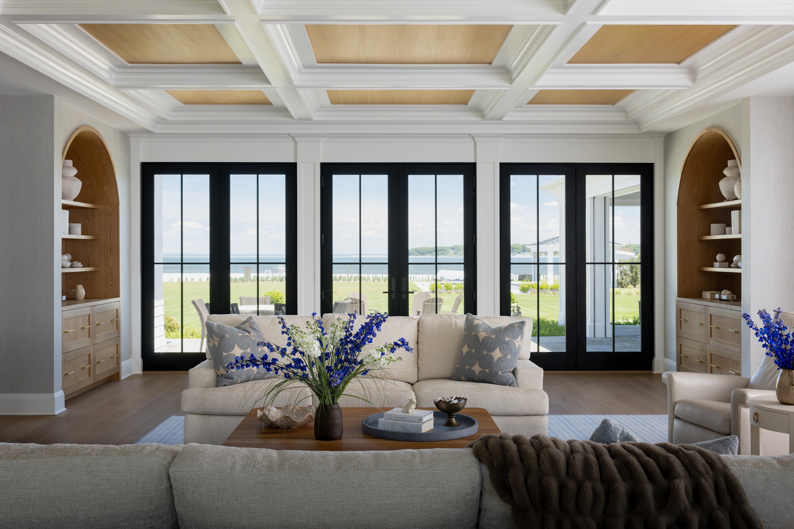 A great room with arched white oak built-ins and a coffered ceiling, with the Long Island Sound visible through black French doors.
