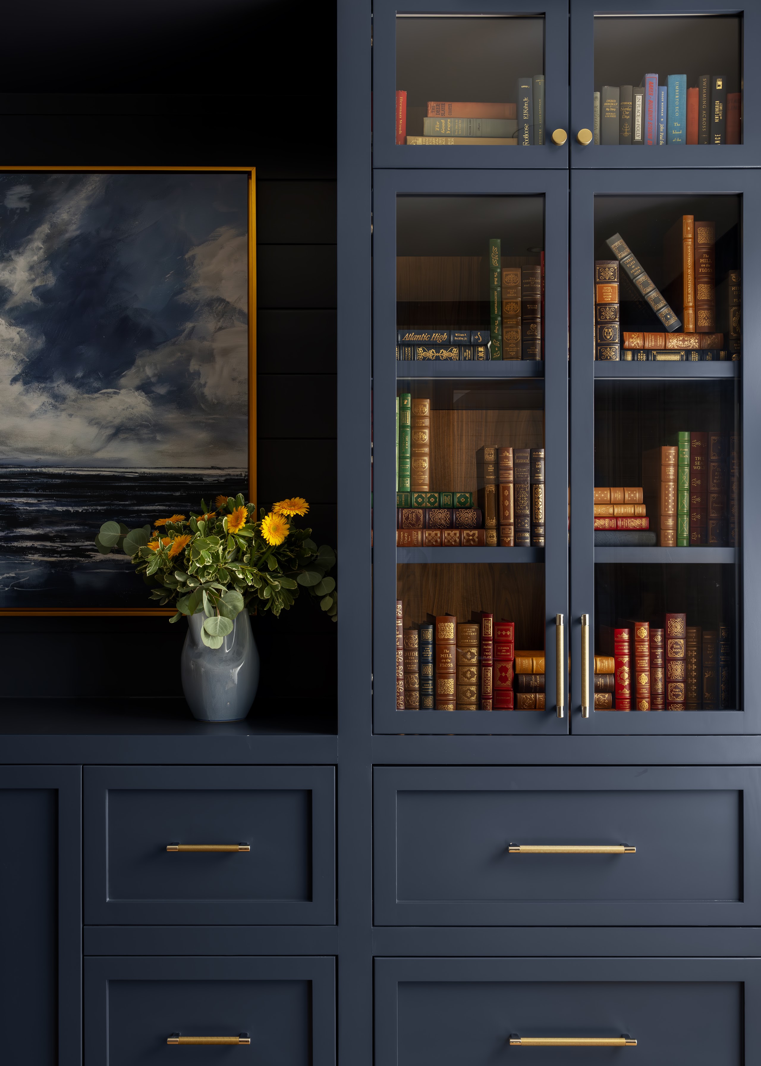 A navy home office with floor-to-ceiling glass-front built-in shelving, leather-bound books, a centered sailboat painting, and yellow flowers.