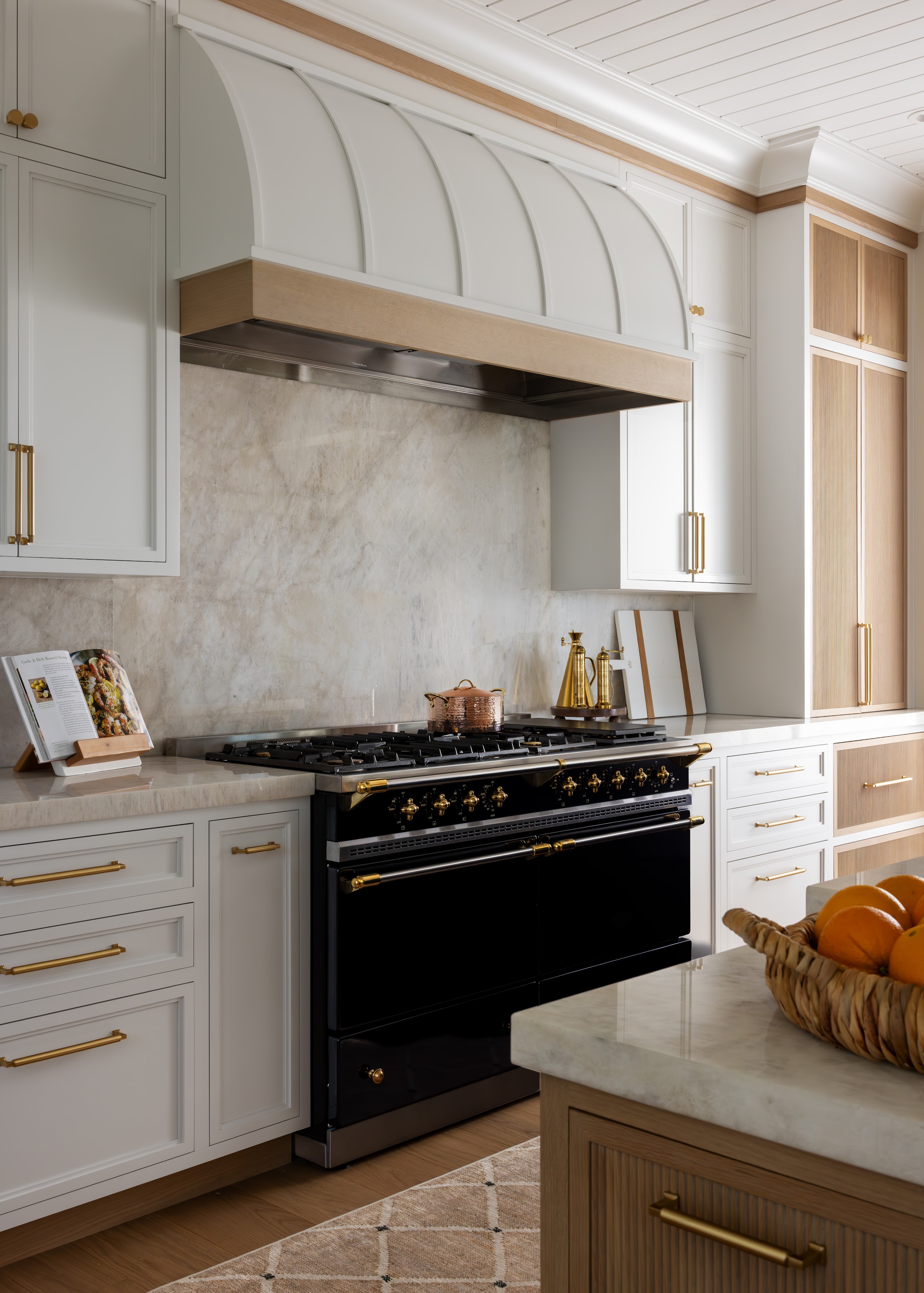A black La Cornue range beneath a custom arched hood, framed by white cabinetry, a natural marble backsplash, and an oak island.