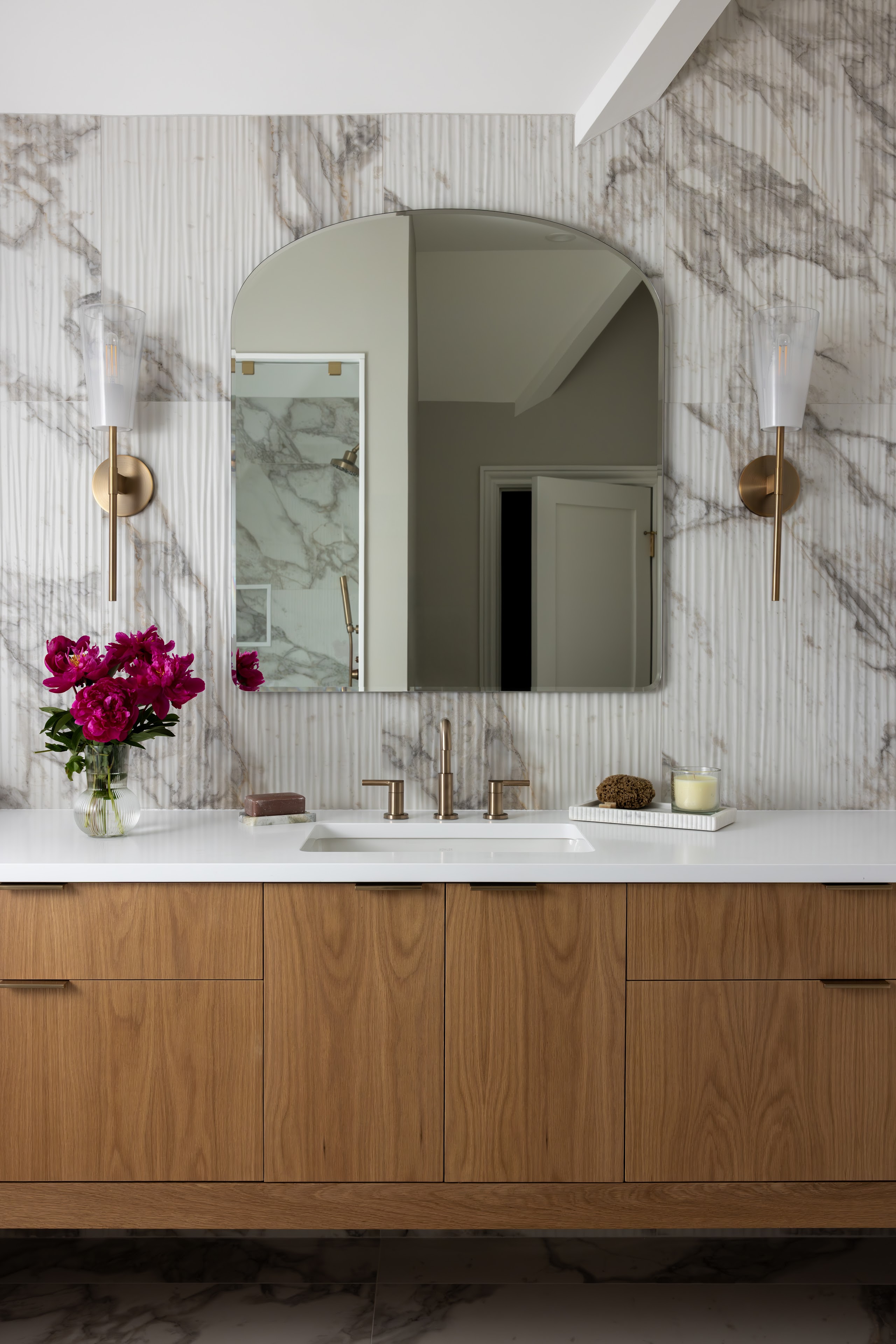A bathroom vanity with a reeded oak cabinet, Cristallo stone backsplash, arched mirror, brass sconces, and hot pink peonies on the counter.