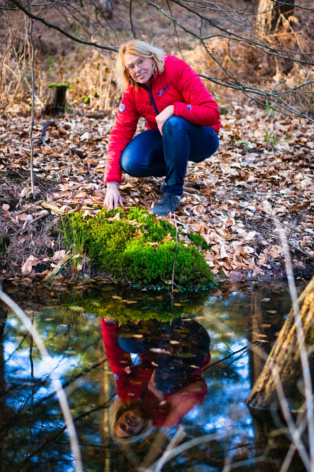 Woman in red jacket crouching by a pond surrounded by dry leaves touching green moss near water