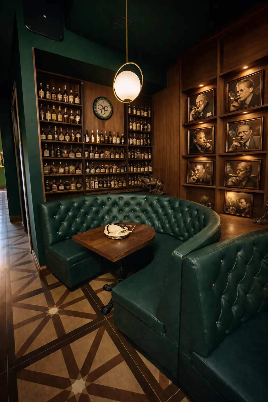 Cozy bar corner with green leather tufted booth seating, wooden table, shelves of liquor bottles, and framed portraits of men smoking cigars on wood-paneled walls.