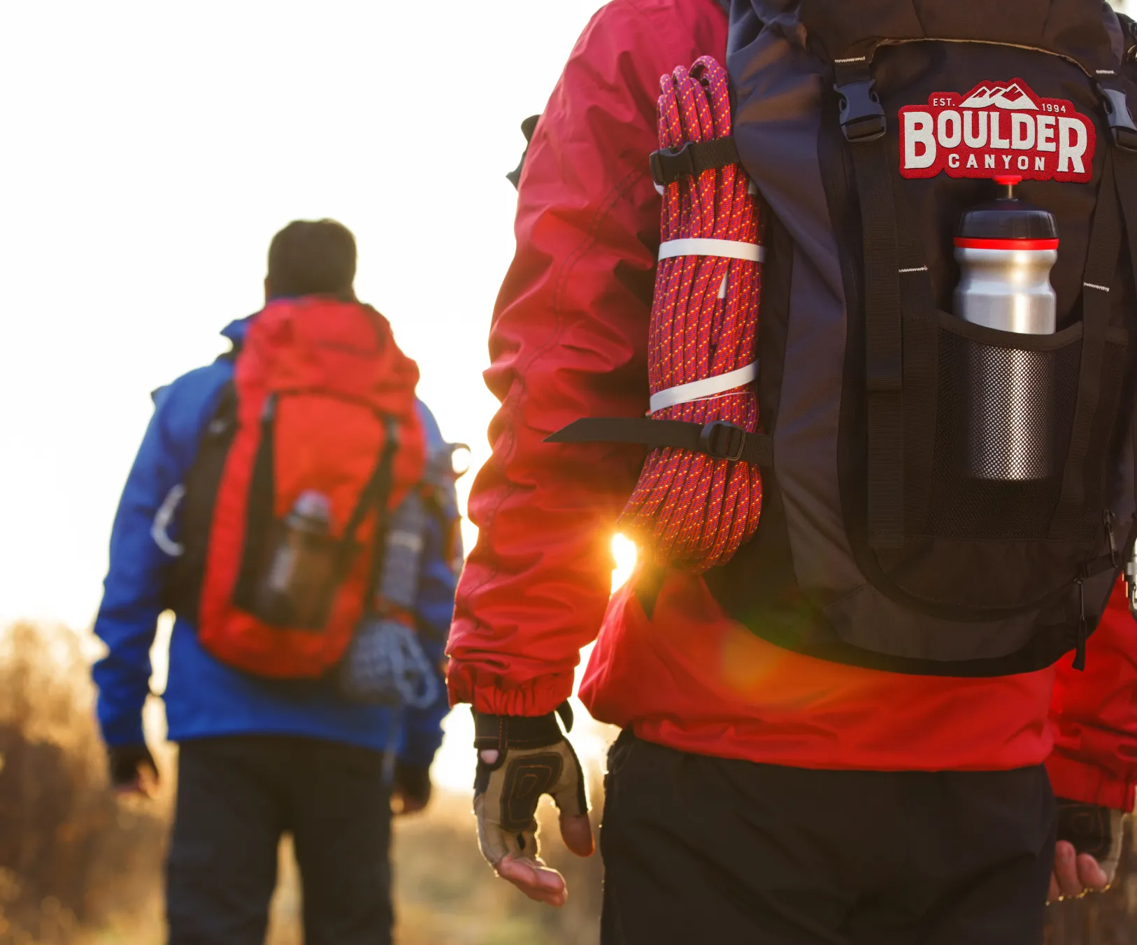 Hikers at sunset, one carrying a backpack with the Boulder Canyon logo patch.