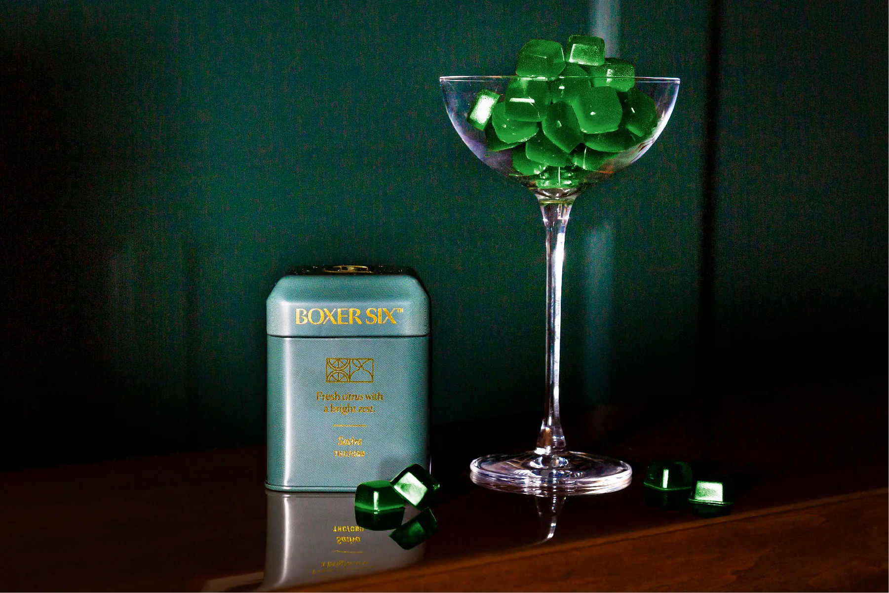 Teal Boxer Six tin beside a coupe glass filled with green diamond-cut gummies
