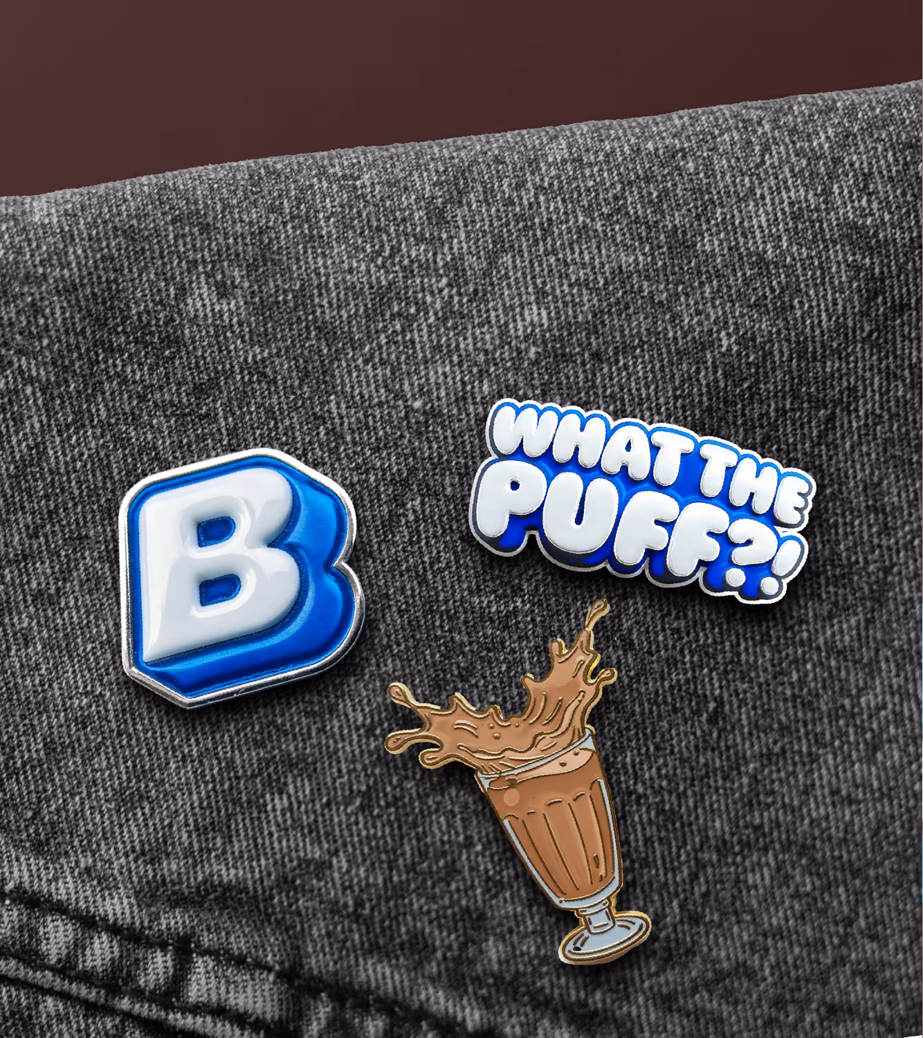 Three Built enamel pins on gray denim: a puffy 'B', a 'WHAT THE PUFF?!' bar, and a chocolate milkshake glass.
