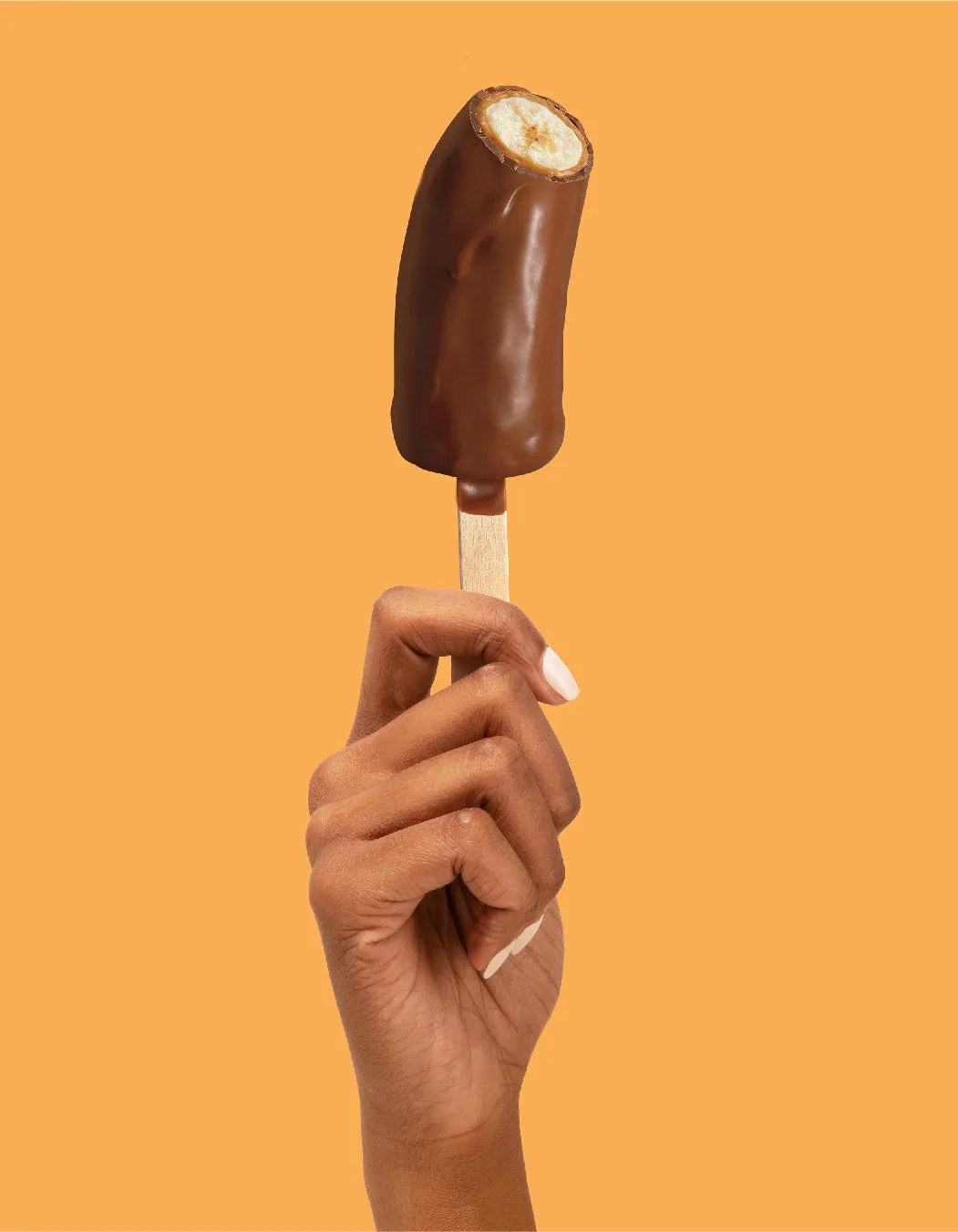 Hand with white nails holding an unwrapped chocolate-dipped frozen banana on a wooden stick on orange.