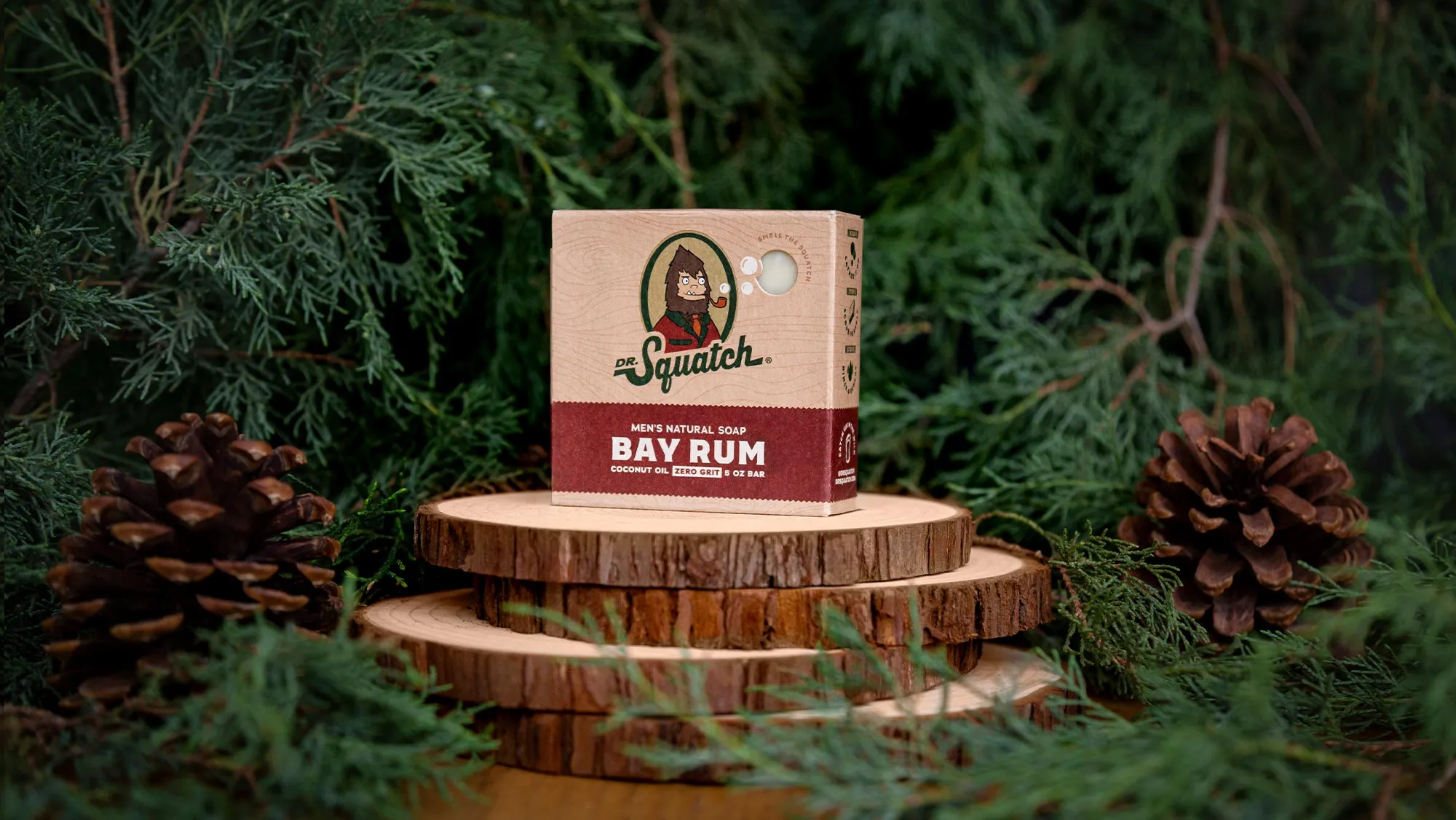 Bay Rum Dr. Squatch soap box on stacked wood slices with cedar foliage and pine cones.