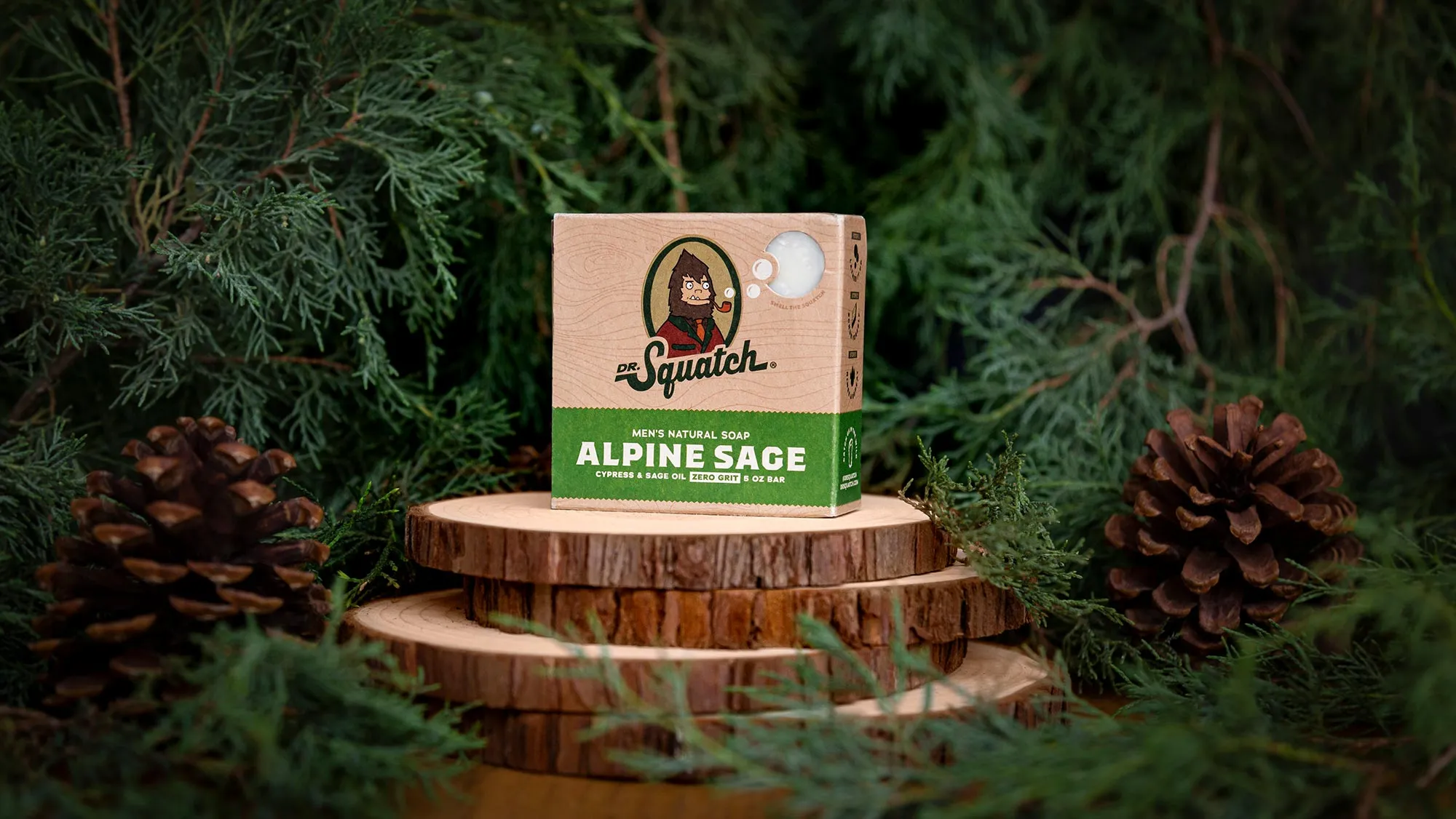 Alpine Sage Dr. Squatch soap box on stacked wood slices with cedar branches and pine cones.