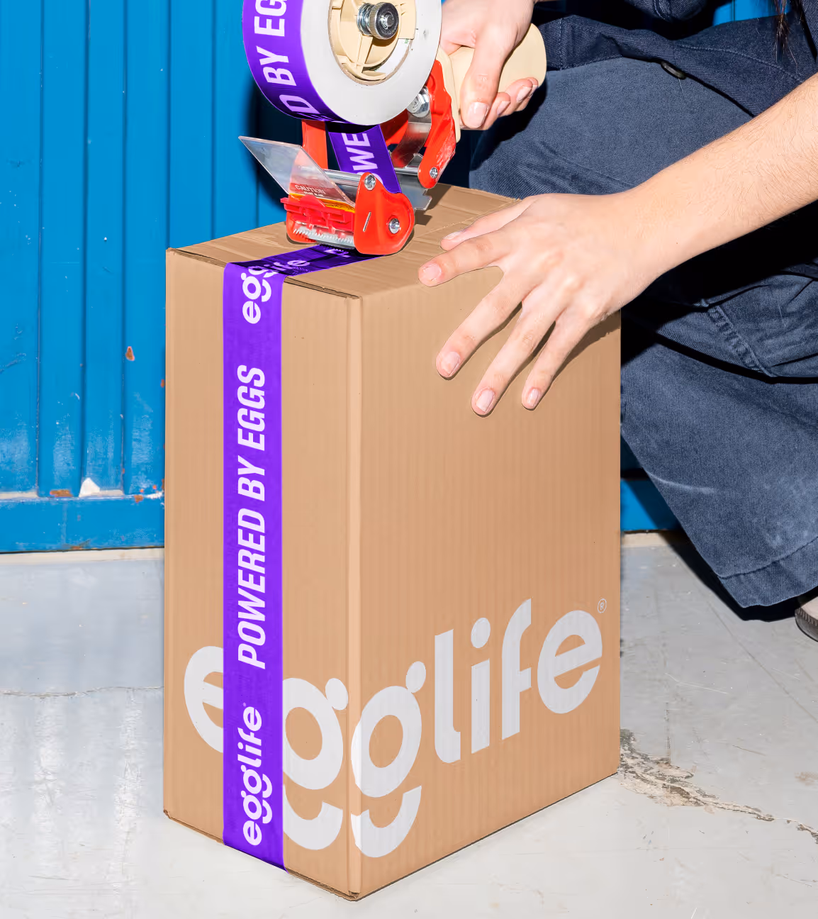 Person sealing a cardboard shipping box with purple 'Powered by eggs' branded tape against a blue garage door.