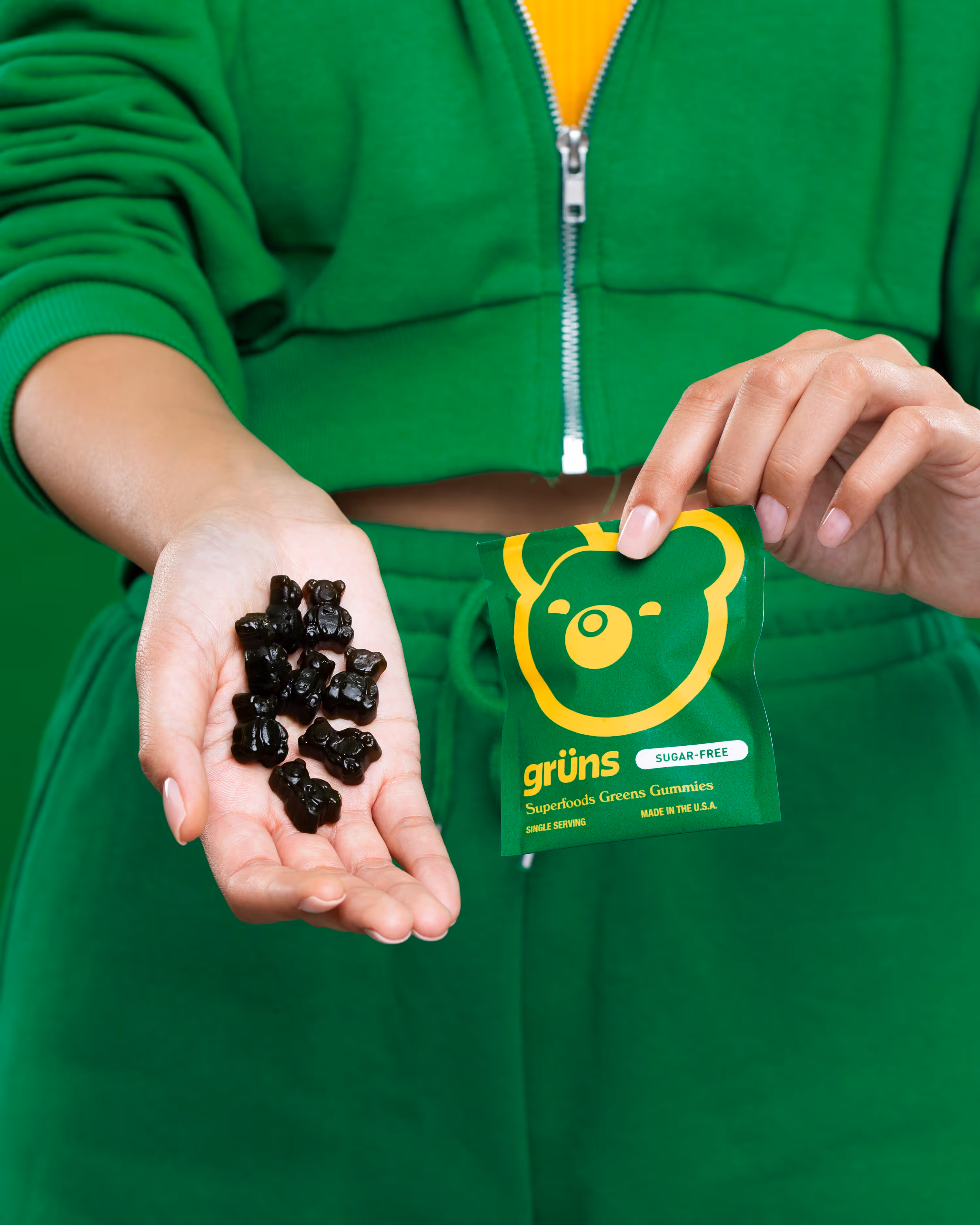 Person in a green hoodie holding dark Grüns gummies and a green Superfoods Greens Gummies sachet with a yellow bear logo.