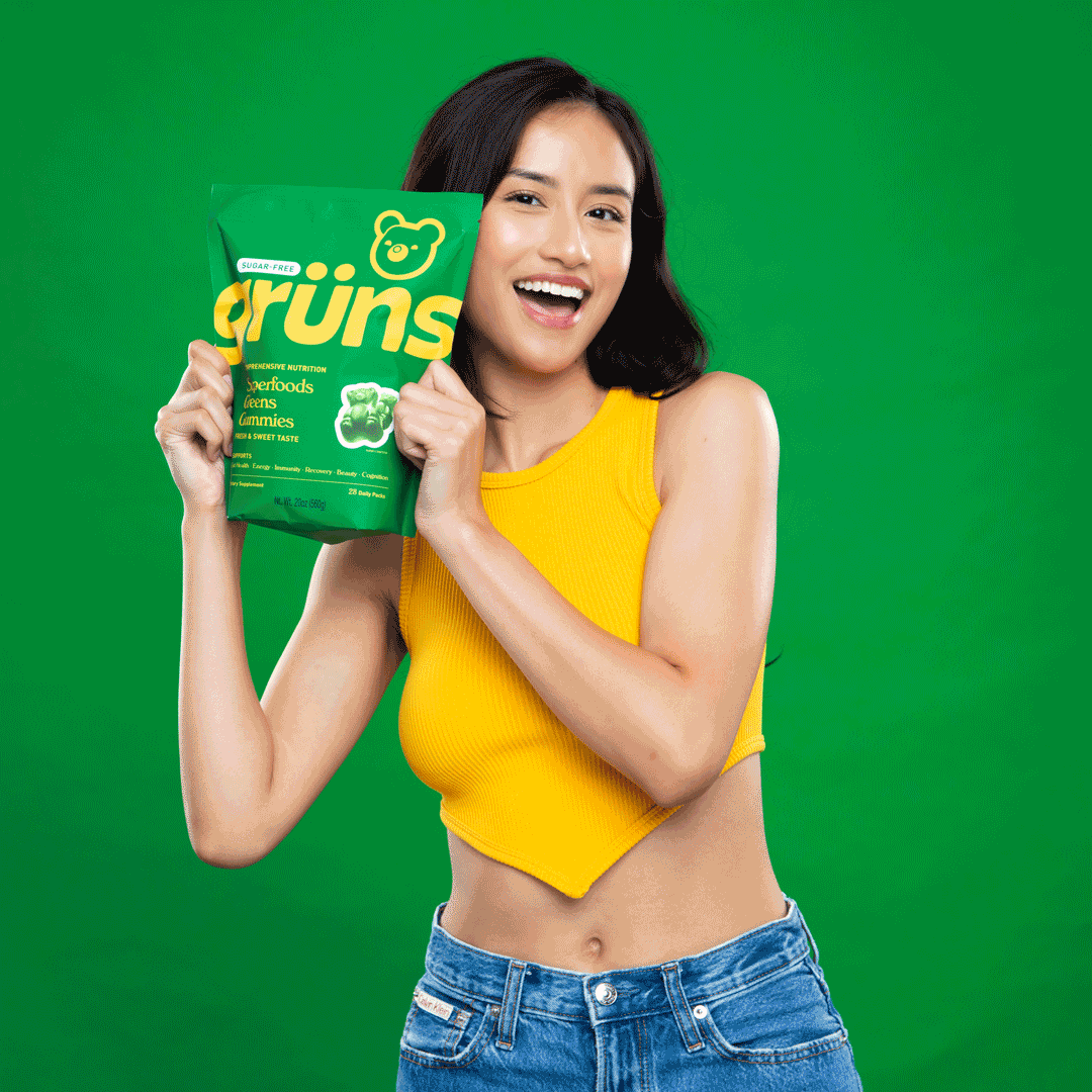 Smiling woman in a yellow tank top on a green backdrop holding a green Grüns Superfoods Greens Gummies pouch.