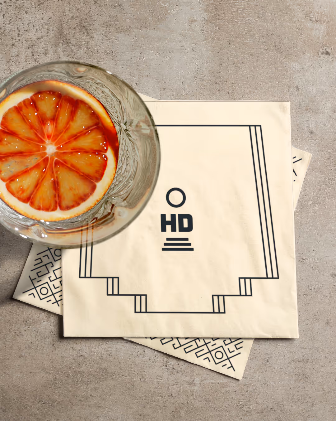 Cream cocktail napkin printed with the stepped 'HD' mark beside a glass with a blood-orange slice on concrete.