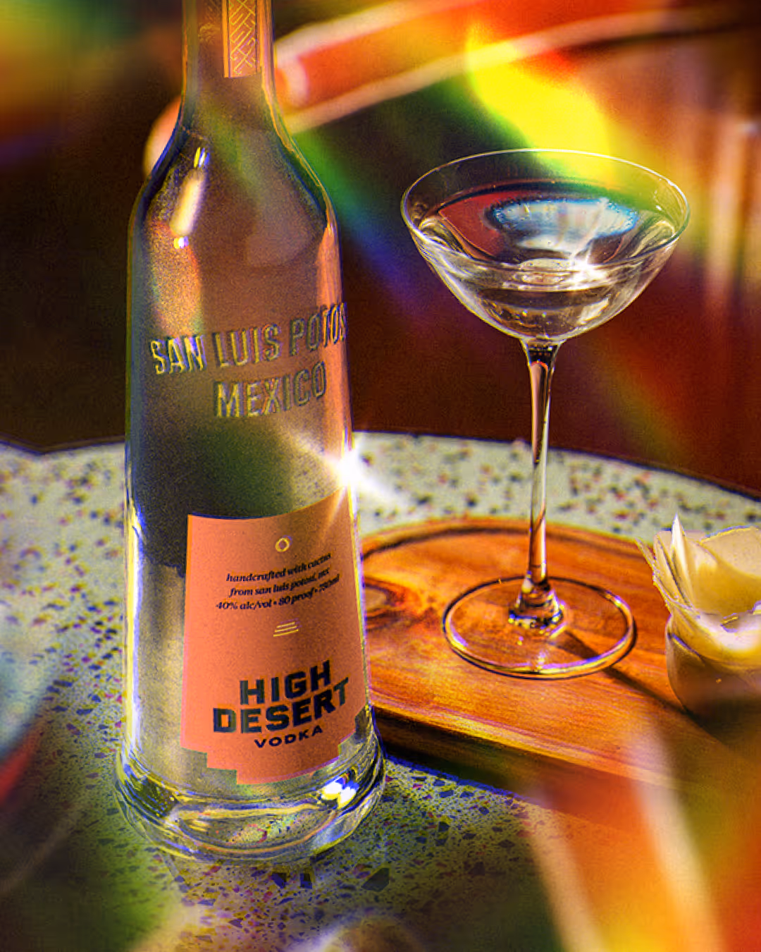High Desert vodka bottle and an empty coupe on a speckled stone tray with prismatic rainbow light streaks.