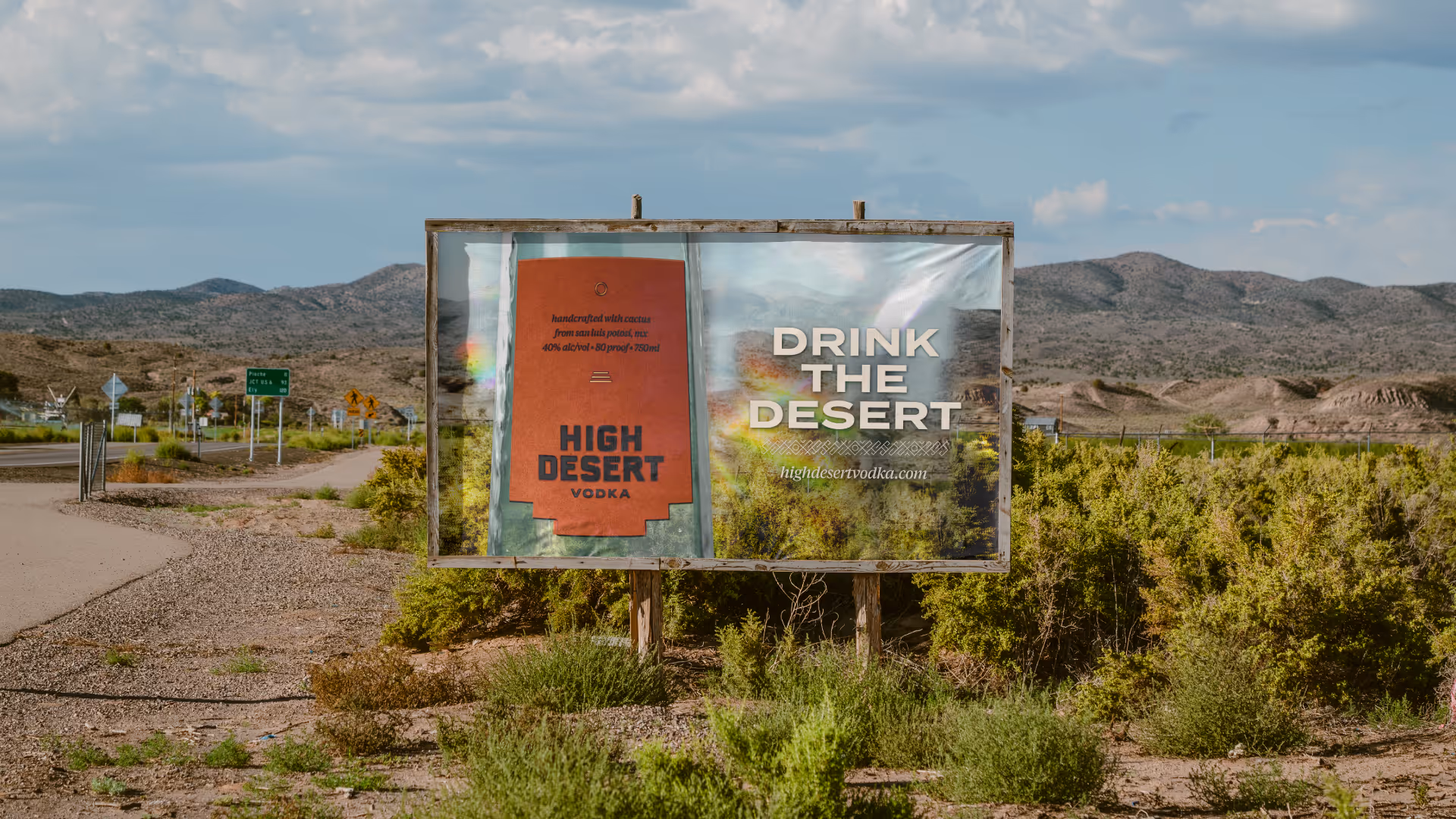 Roadside billboard in arid mountains reading 'Drink the Desert — highdesertvodka.com' beside an orange brand panel.
