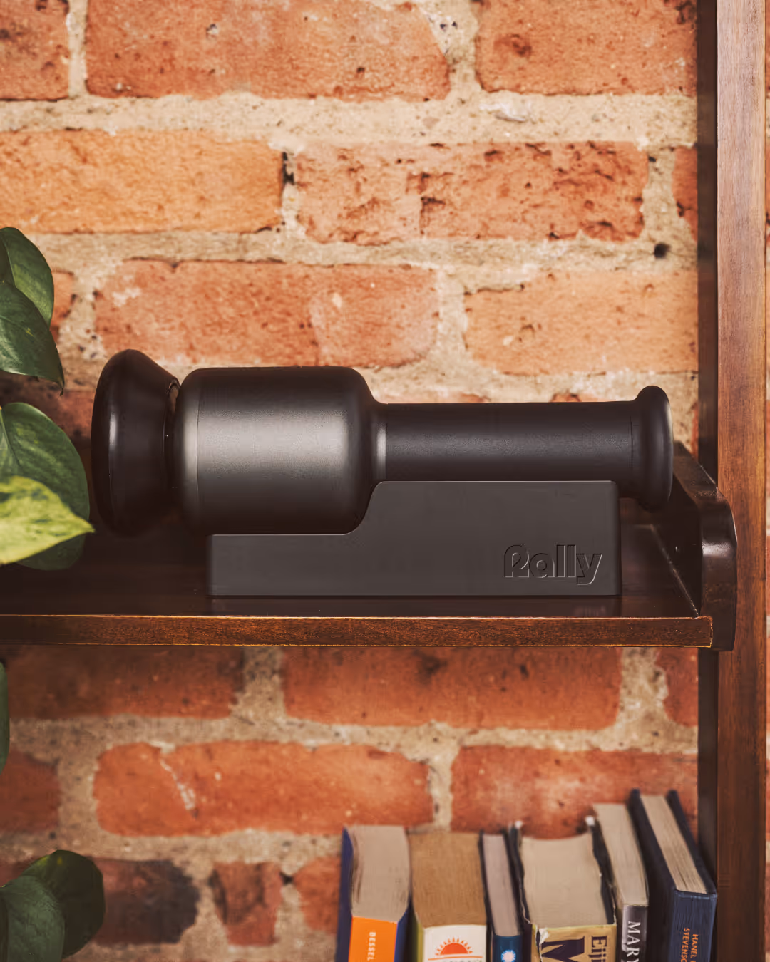 Black Rally massage device docked in its charging base on a wooden shelf against exposed brick with plants and books.