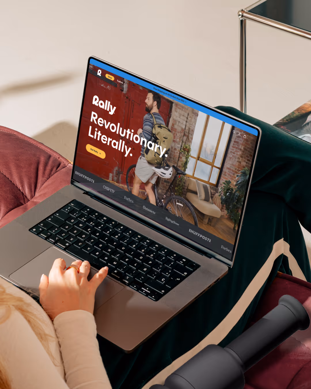 Laptop on a woman's lap showing the Rally homepage with 'Revolutionary. Literally.' headline and a cyclist photo.