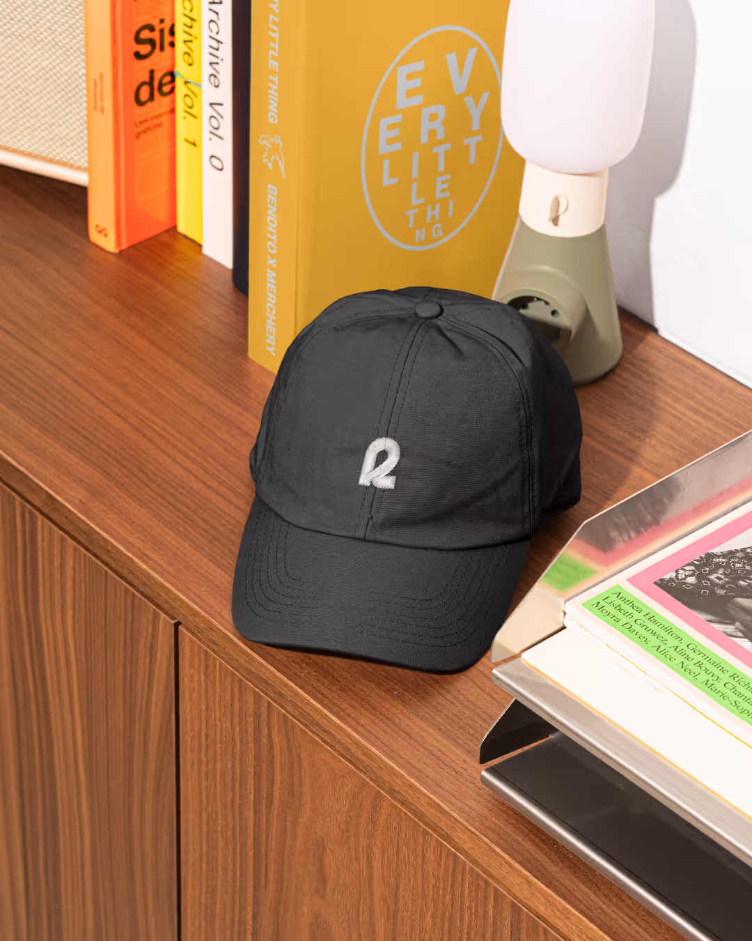 Black Rally baseball cap with embroidered white 'R' on a wooden console with books, lamp, and a yellow poster.