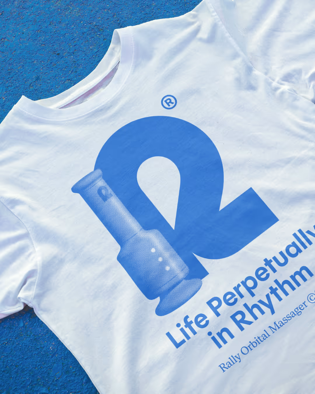 White Rally tee on blue speckled surface printed with a blue 'R' logo and 'Life perpetually in rhythm' tagline.
