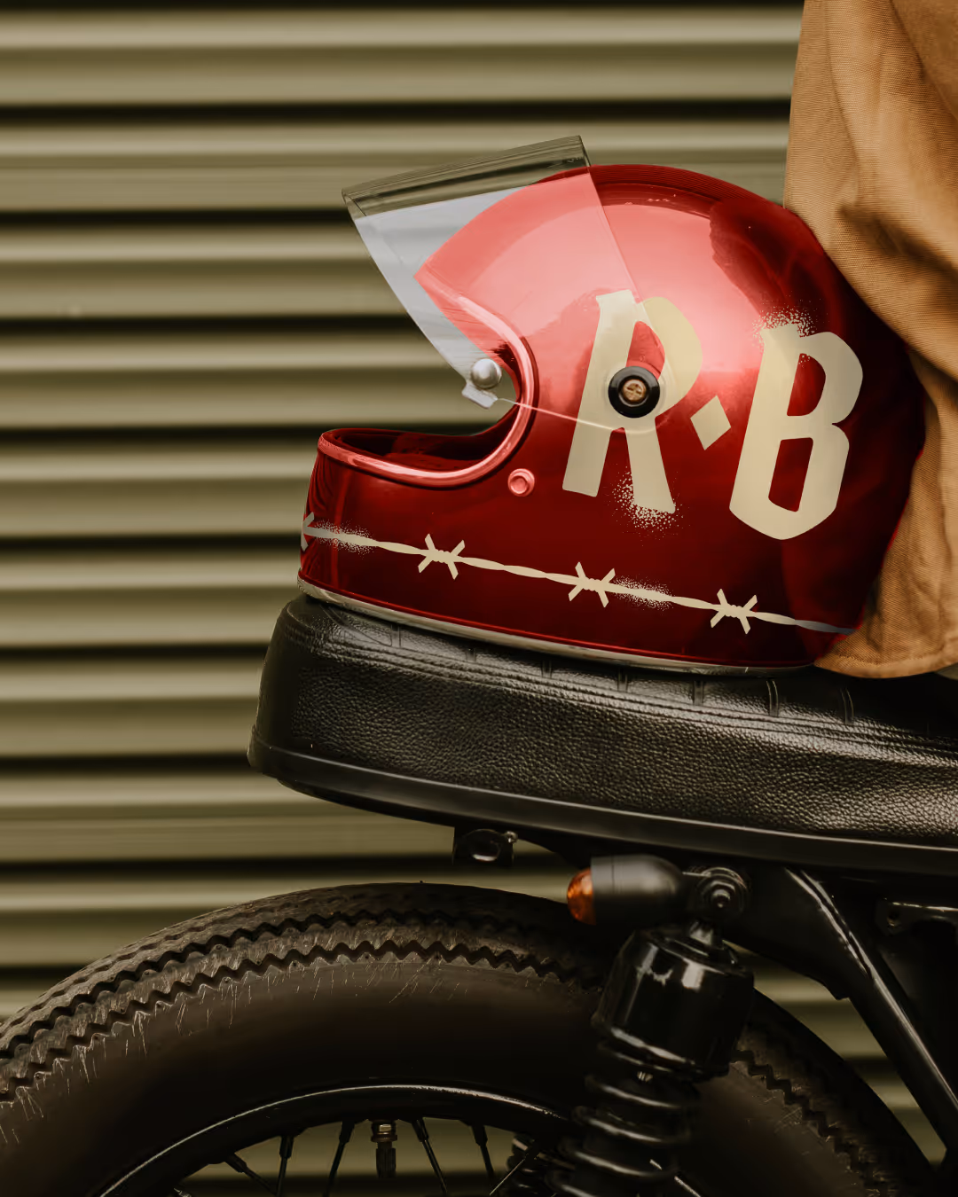 Glossy red Rare Breed motorcycle helmet with cream 'R-B' lettering and barbed-wire graphic on a cafe-racer seat.