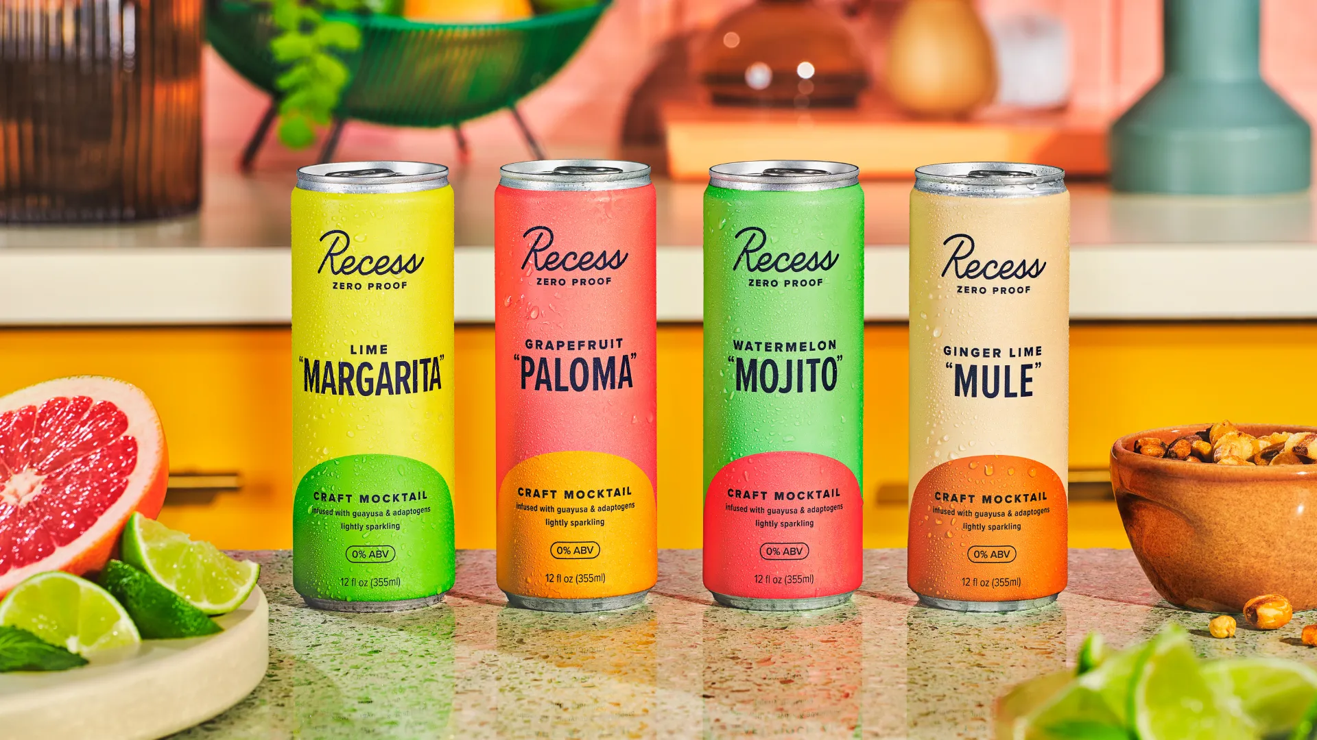 Four Recess Zero Proof cans — Lime Margarita, Grapefruit Paloma, Watermelon Mojito, Ginger Lime Mule — on a counter with citrus fruits and herbs.