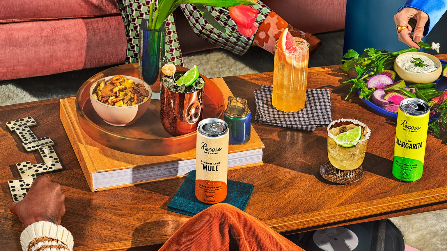 Overhead party table with Recess Zero Proof Ginger Lime Mule and Lime Margarita cans among dominoes, books, snacks, and mixed drinks.