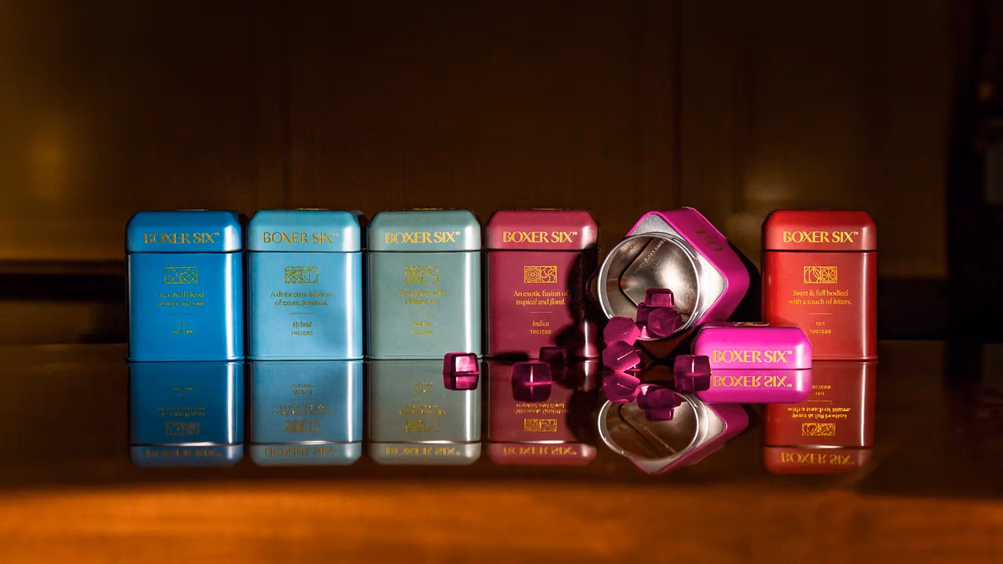 Six Boxer Six tins in gradient colors from teal to red line up on a dark surface, one open to reveal red gummies inside.