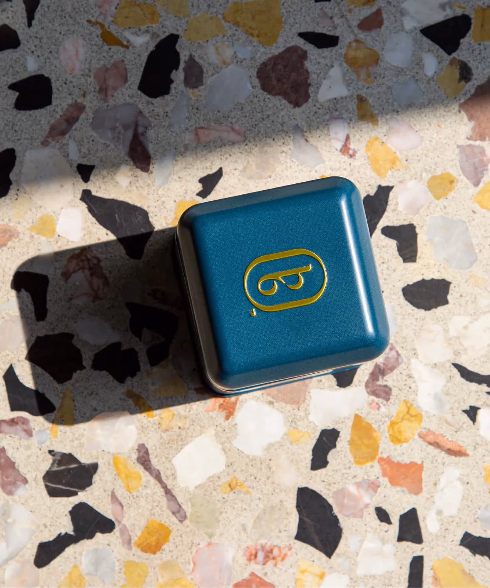 A square teal Boxer Six tin with a gold logo rests on a terrazzo surface in bright sunlight.