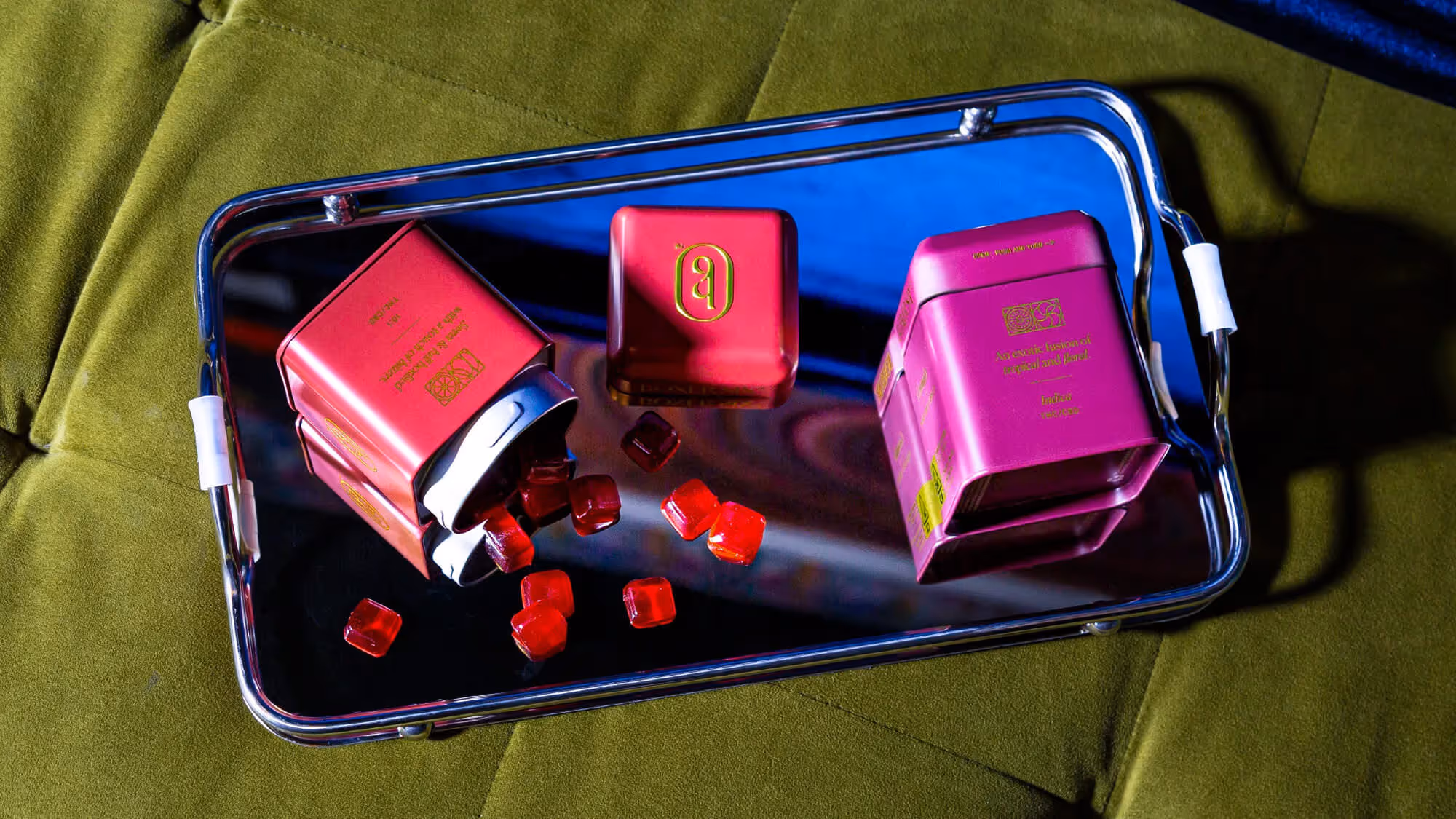 Two magenta Boxer Six tins and scattered red gummies on a mirrored blue tray over a green textured surface.