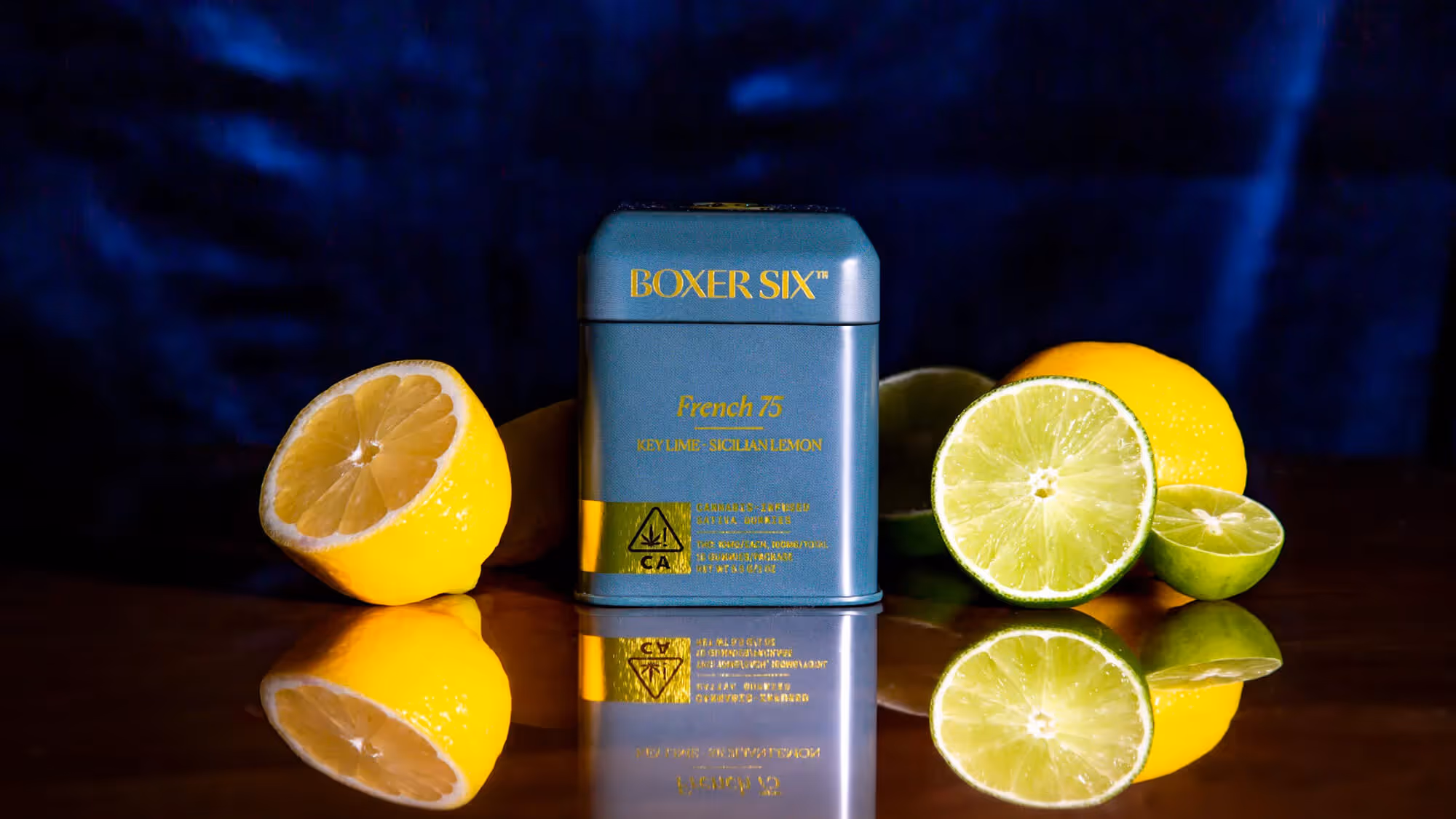 A teal Boxer Six 'French 75' tin surrounded by lemons and lime halves on a dark surface with blue velvet behind.
