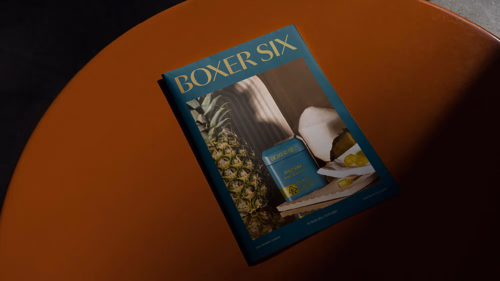 A Boxer Six branded magazine rests on an orange curved surface, its cover showing a pineapple and teal tin.