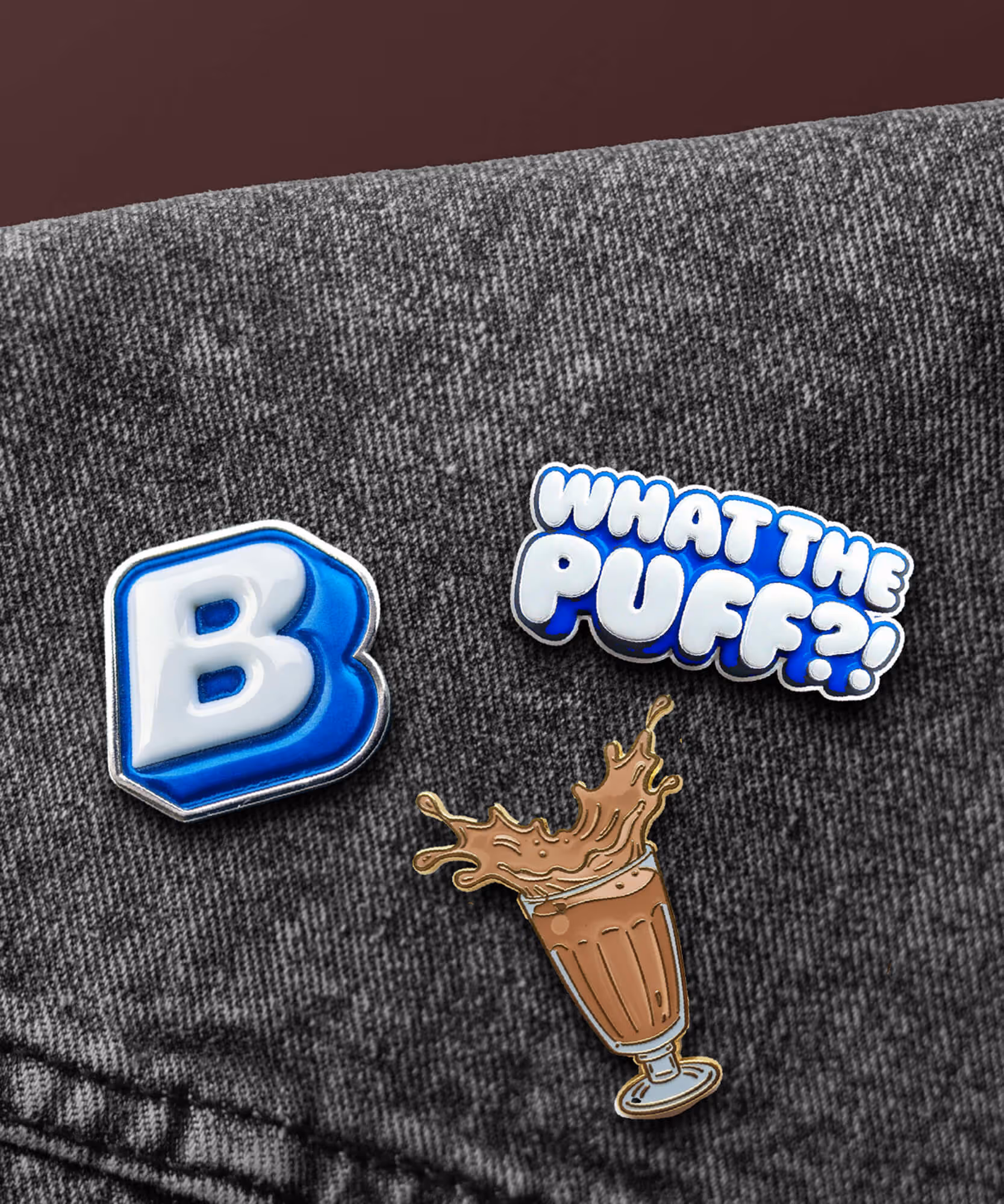 Three Built enamel pins on gray denim: a puffy 'B', a 'WHAT THE PUFF?!' bar, and a chocolate milkshake glass.