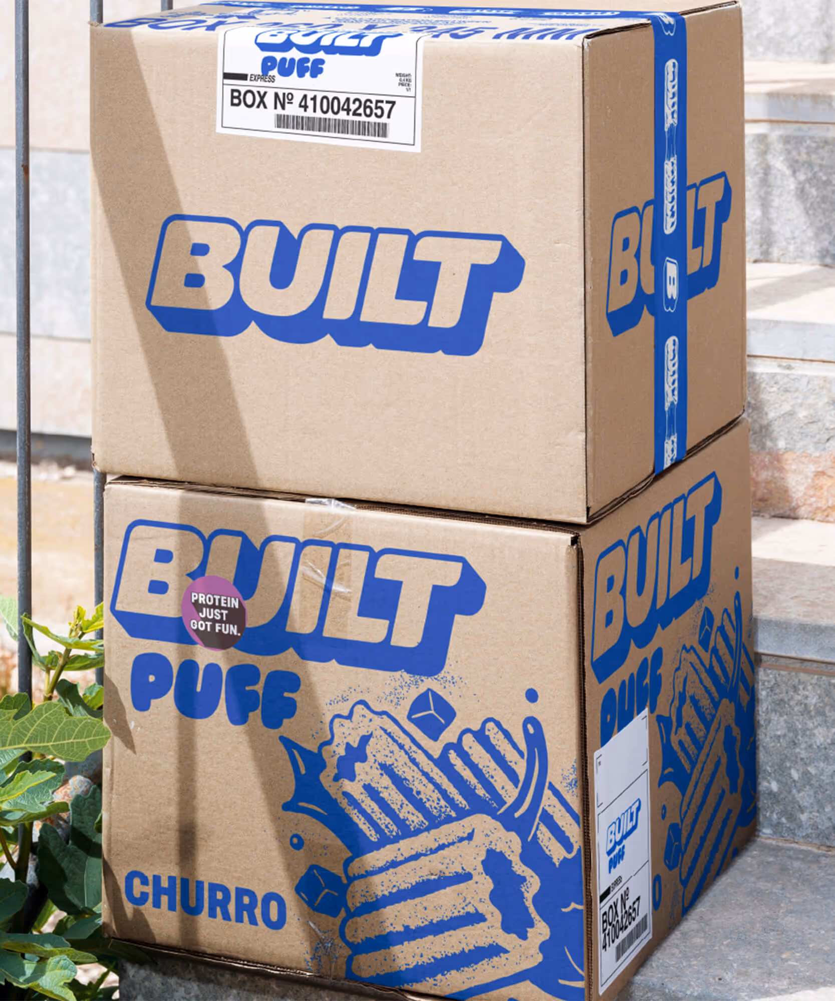 Two cardboard shipping cartons printed with blue Built and Built Puff Churro logos on a concrete step.