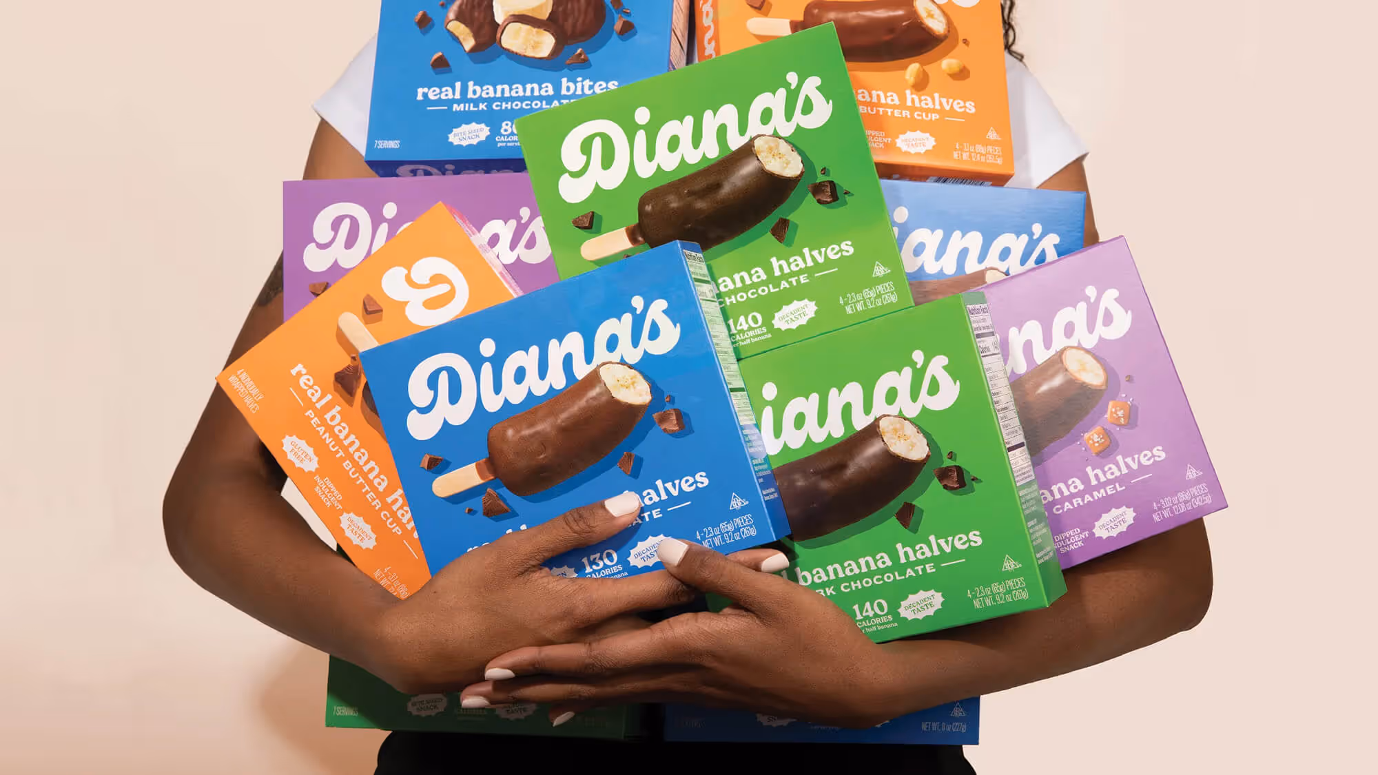 Person hugging a stack of Diana's boxes in green, purple, blue, and orange against a beige wall.